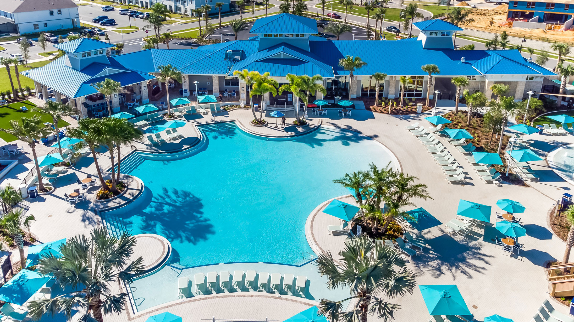 An aerial view showcases a large, irregularly shaped communal pool with bright blue water, surrounded by numerous lounge chairs and teal umbrellas. A large clubhouse with a blue roof and palm trees completes the resort-like setting.