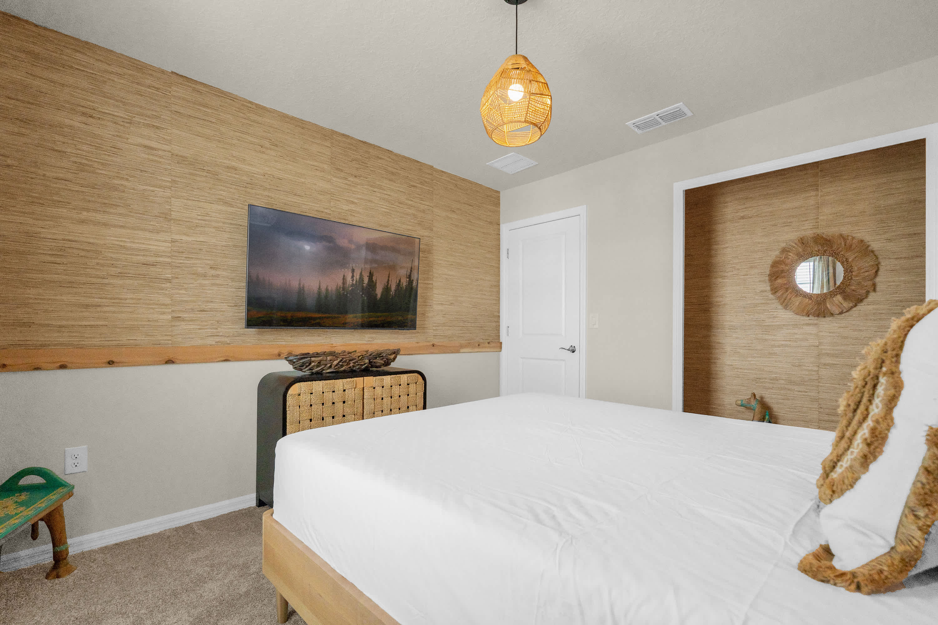 Another view of the tropical-themed bedroom, showing a wall-mounted television above a rattan and wood dresser, with a round mirror and a small green stool in the corner, all against a textured accent wall.