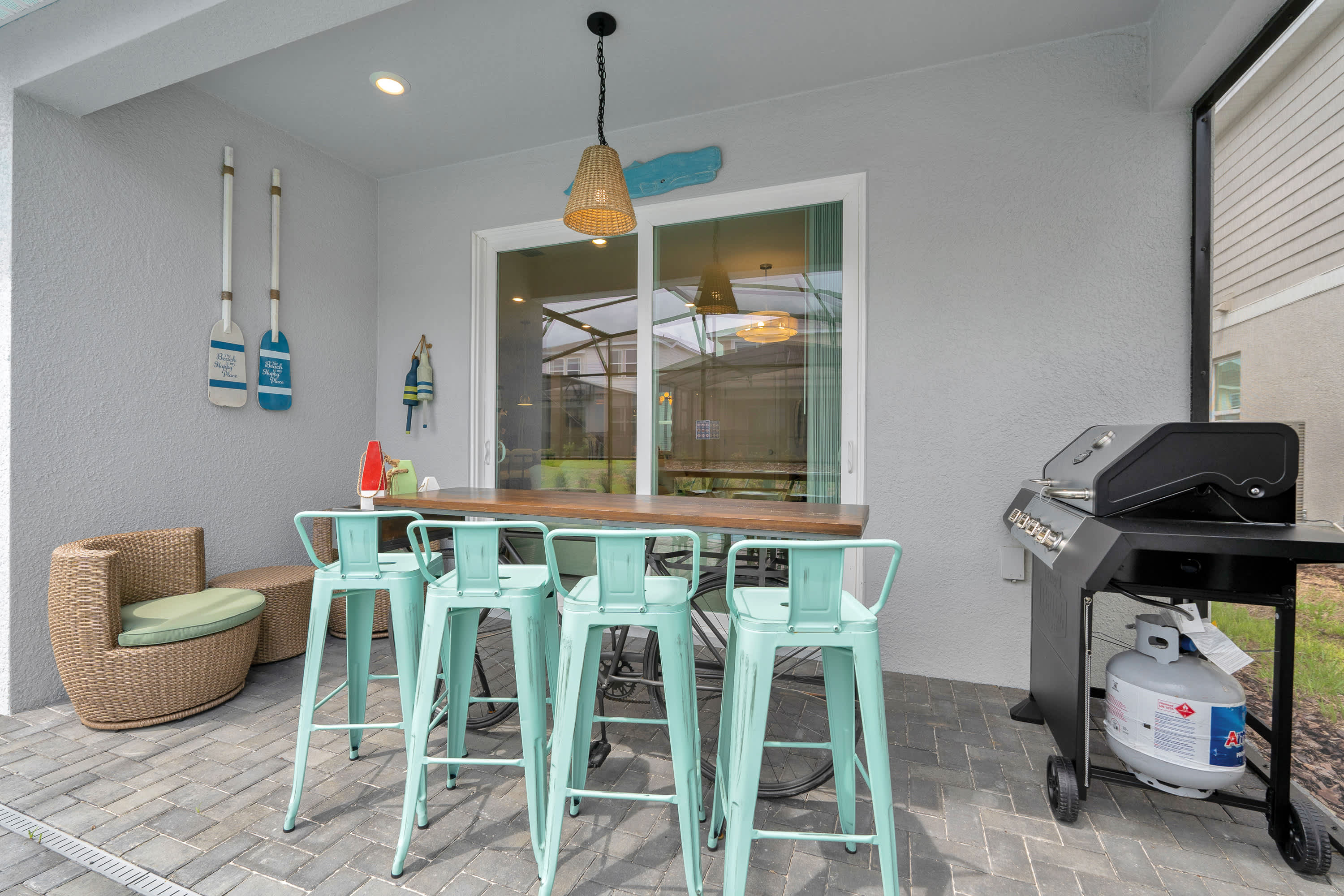 A covered outdoor patio area featuring a long bar table with four teal bar stools and a BBQ grill, decorated with nautical-themed wall decor.