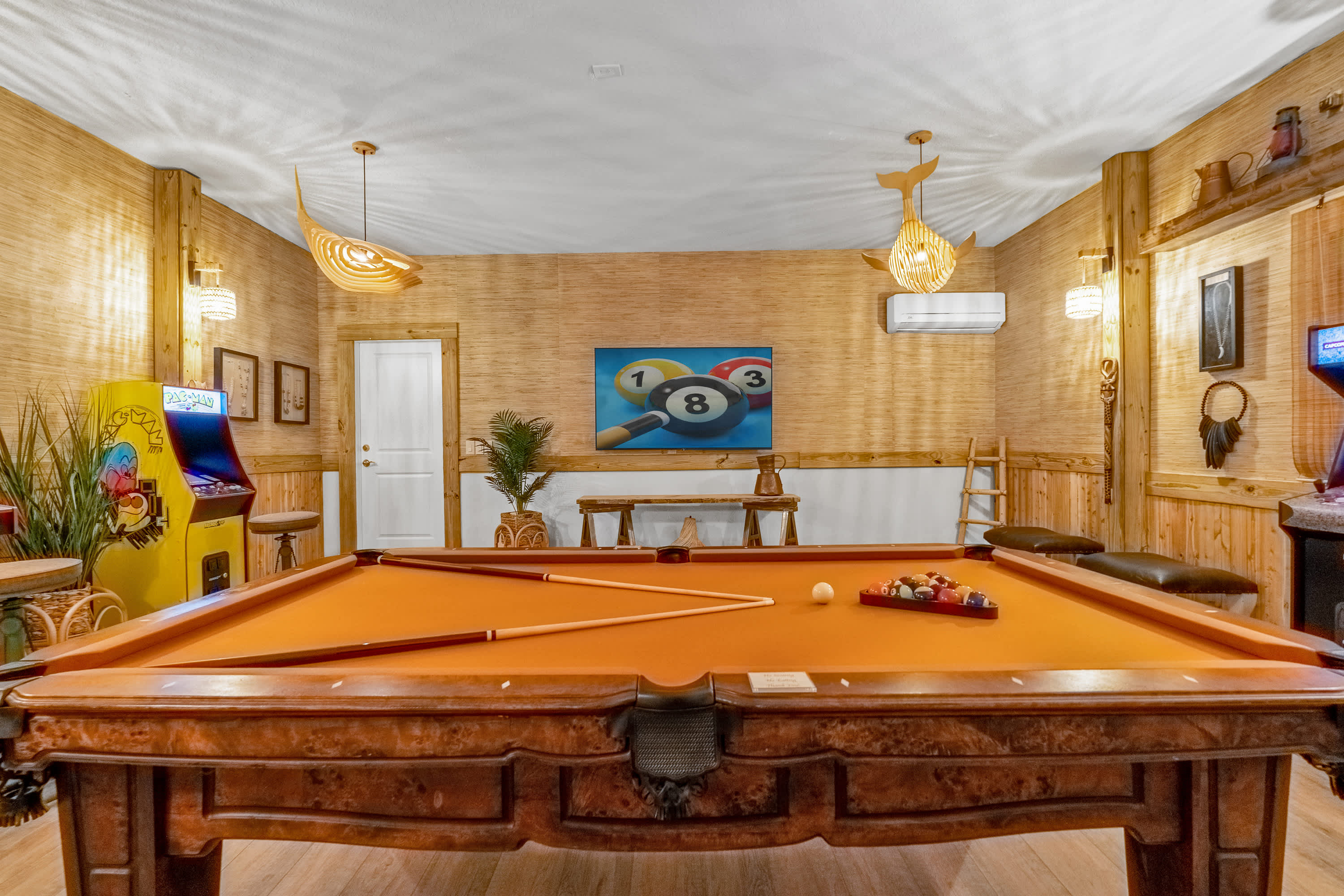 A wide view of a game room centered around a wooden pool table with orange felt, surrounded by walls featuring light wood paneling and woven textures. Two unique fish-shaped pendant lights hang from the ceiling, and a Pac-Man arcade machine is visible.