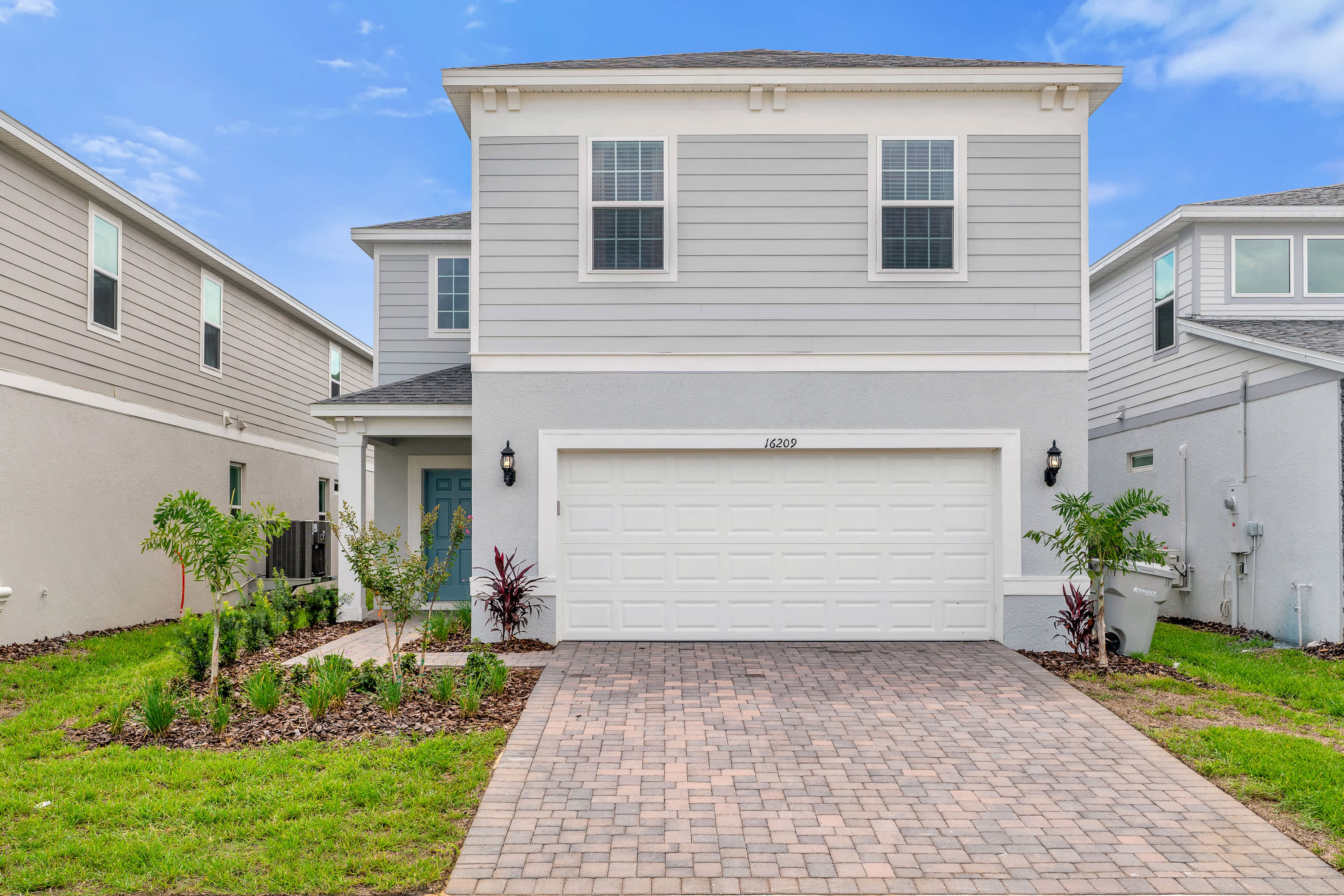 The front exterior of a modern two-story house with light gray siding and a white garage door, featuring a paved driveway and landscaped garden beds leading to a blue front door.
