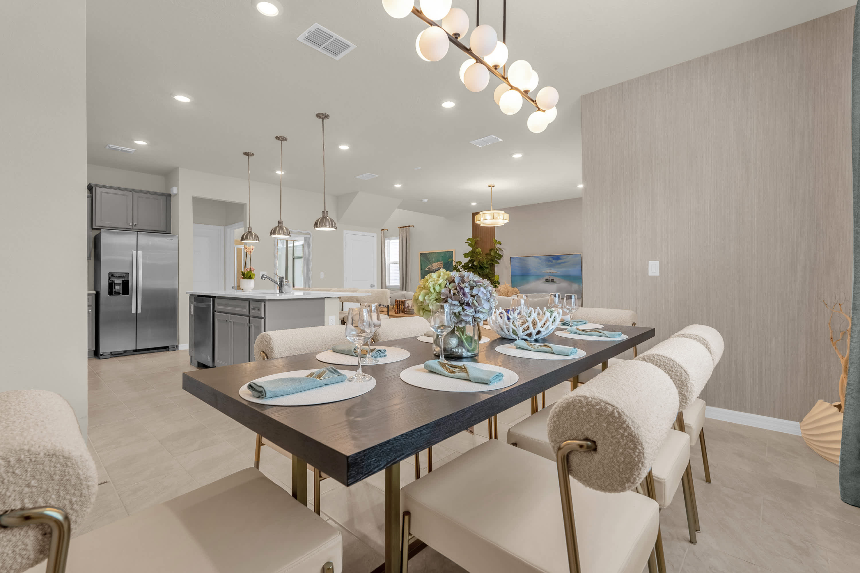 A modern dining area featuring a long dark wood table set with light blue placemats and white chairs with textured backs. A linear chandelier hangs above, and the open-concept kitchen with an island and bar stools is visible in the background.