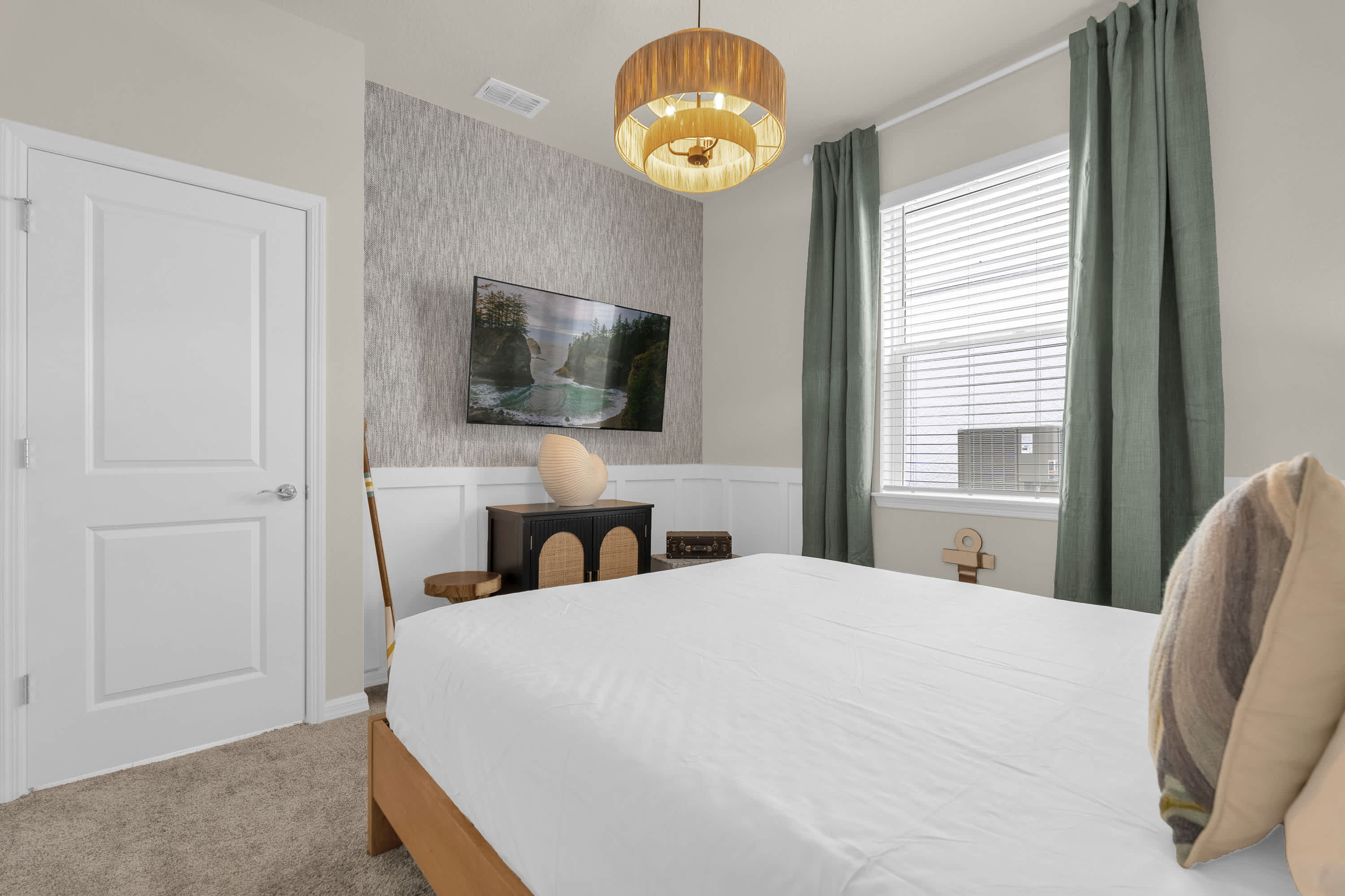This view of the bedroom shows a wall-mounted TV above a dark cabinet, with a decorative vase. The room maintains its coastal theme with wainscoting and a textured accent wall.