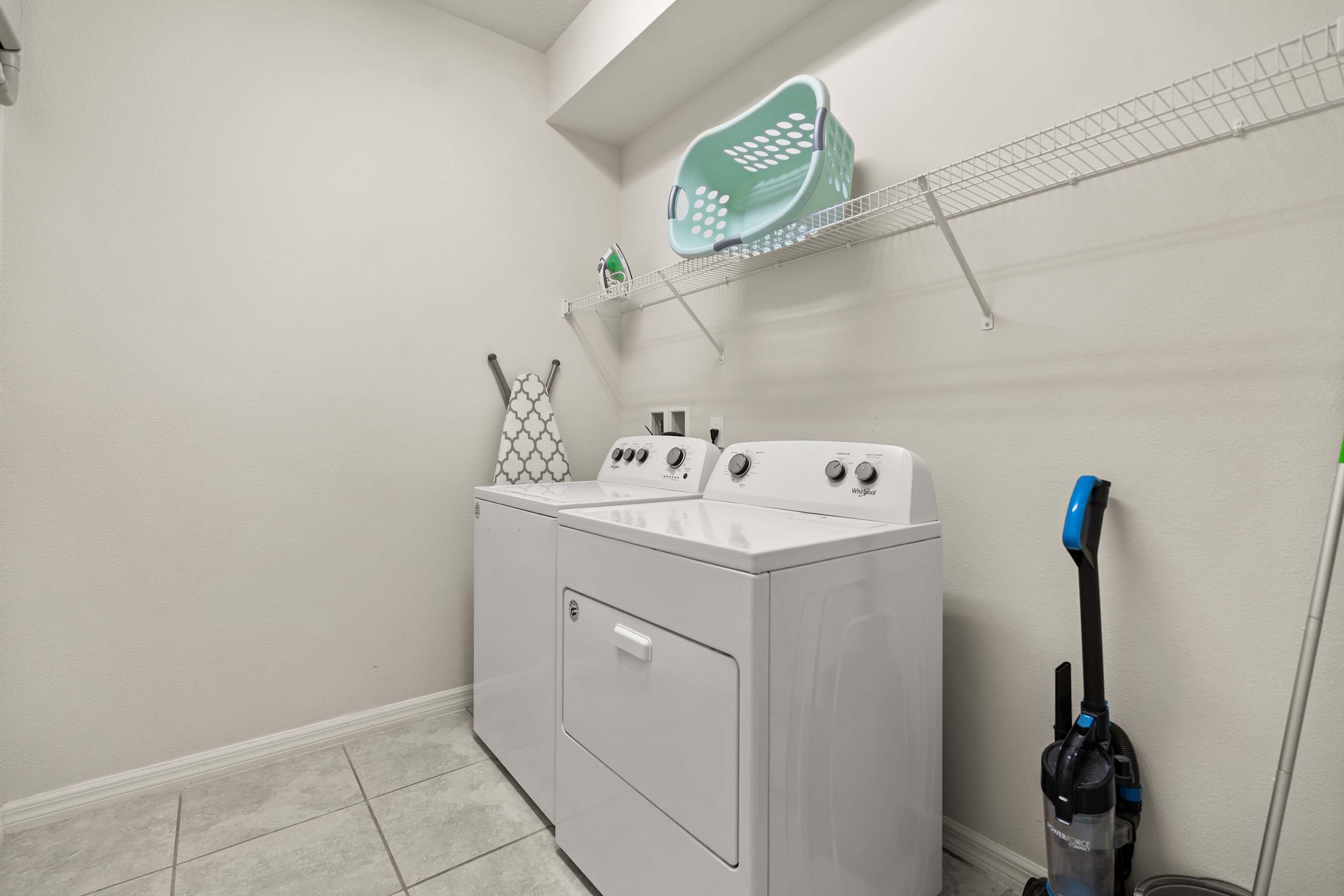 A functional laundry room contains a white top-loading washing machine and a front-loading dryer, with a wire shelf above holding a laundry basket and supplies.
