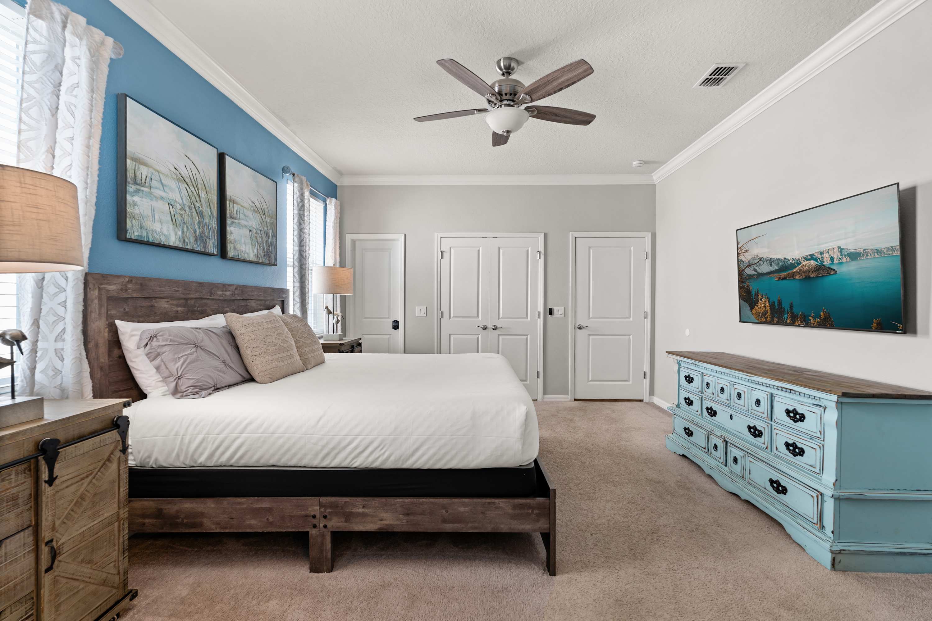 A wide shot of the bedroom with a large wooden king bed, light blue accent walls, and a ceiling fan. A light blue distressed dresser with a TV is on the right, and two white doors lead to closets or another room.