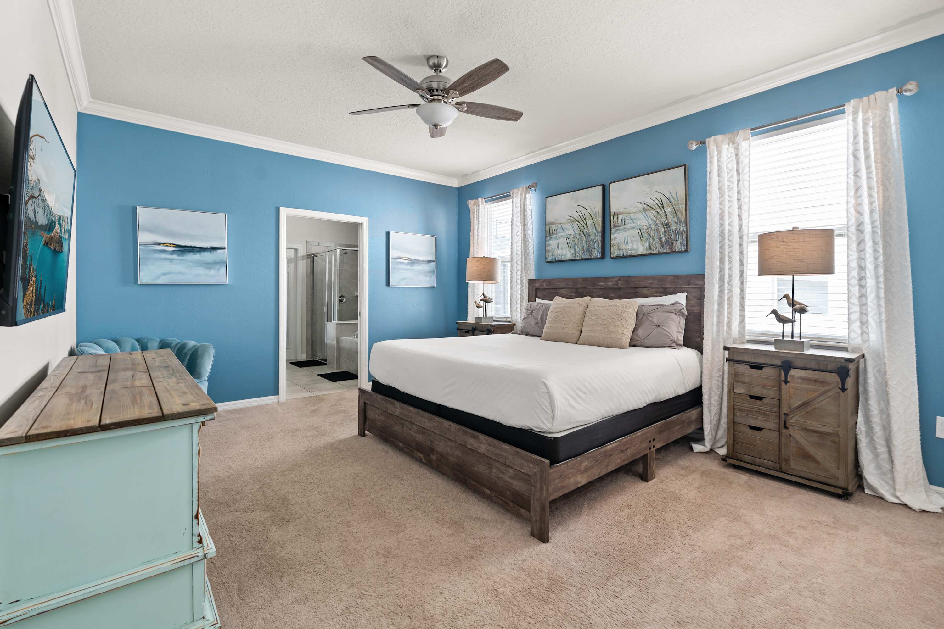 A bright bedroom with vibrant blue accent walls and a large wooden king-sized bed with a dark headboard. Two rustic nightstands with lamps flank the bed, and a light blue dresser with a TV is on the left, with a bathroom entrance visible.