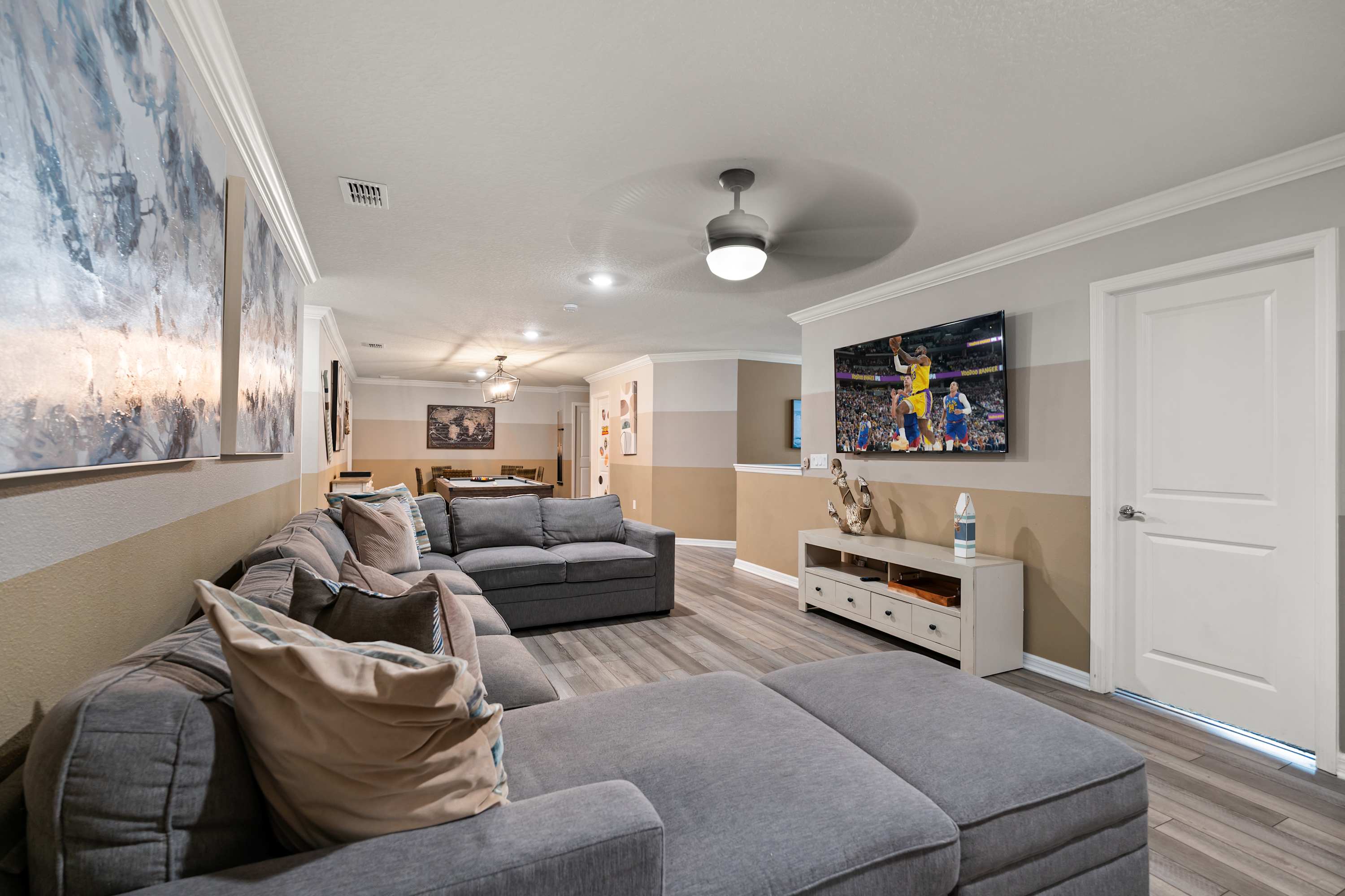 Cozy living room with a large gray sectional sofa, a wall-mounted TV displaying a basketball game, and two-tone walls in beige and light gray, set on light wood-look flooring.