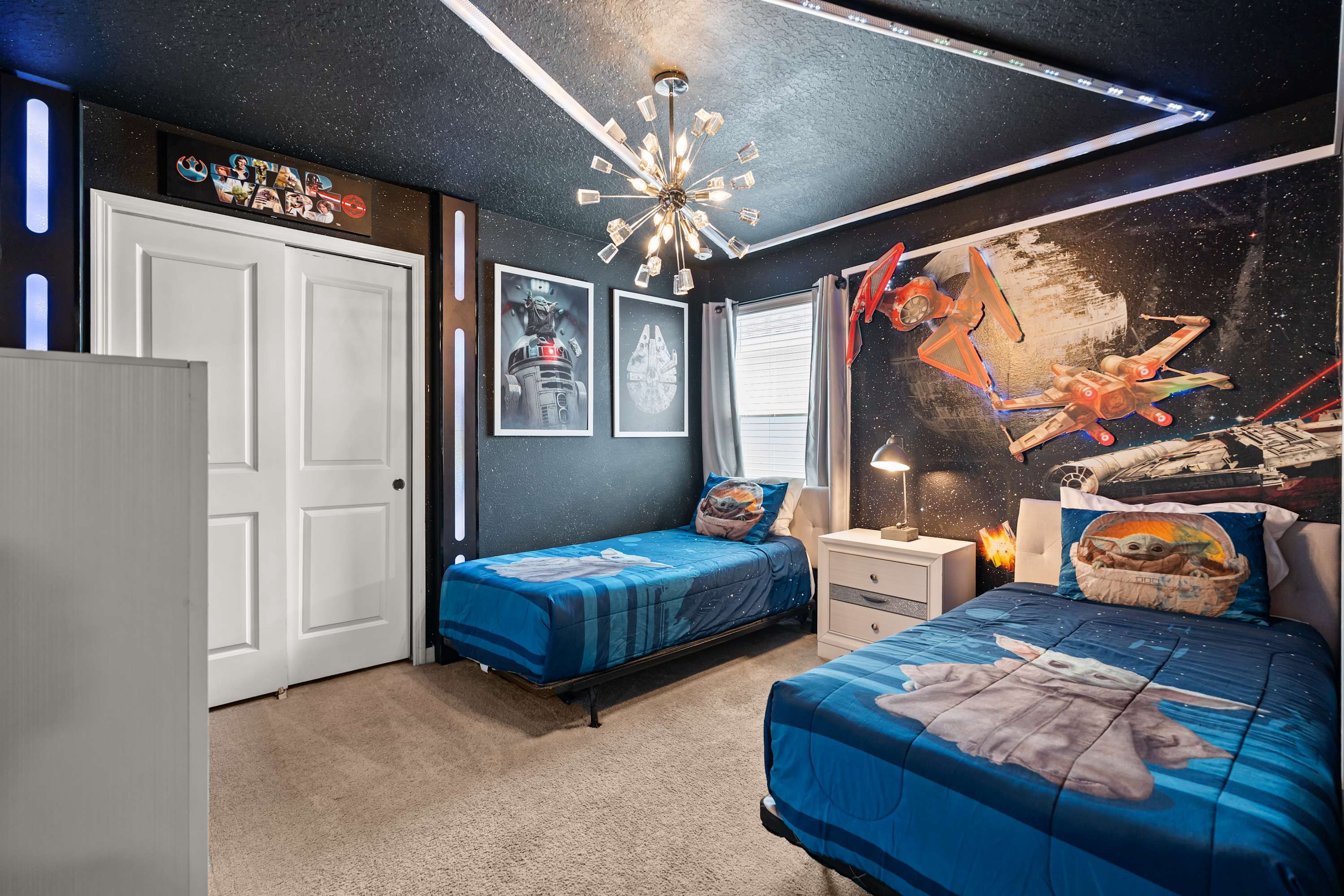 A Star Wars-themed bedroom features two twin beds with Grogu bedding, a dark ceiling with star-like lights, and a wall mural depicting spaceships and characters, illuminated by a modern sputnik chandelier.