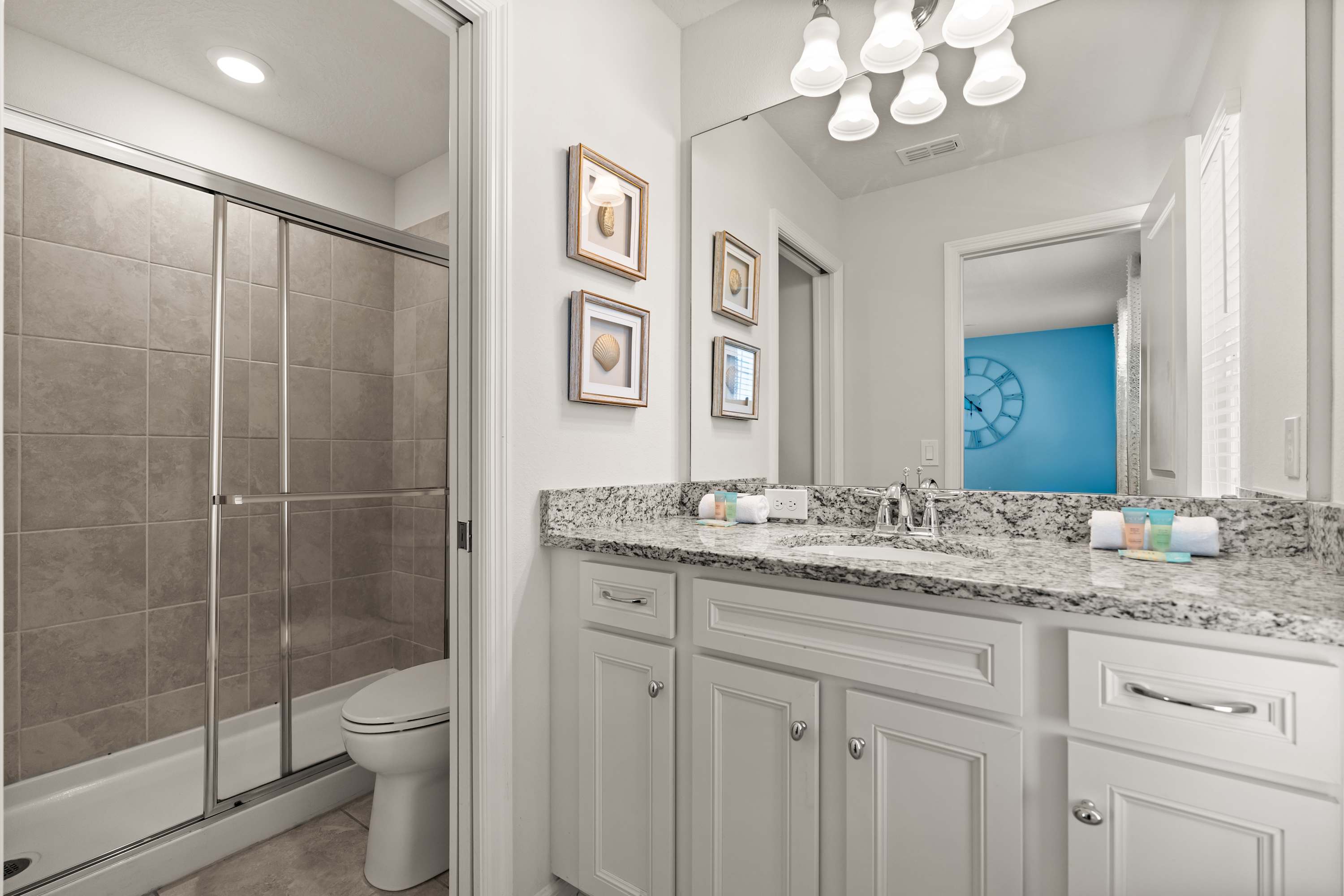 A bright bathroom featuring a white vanity with a speckled granite countertop, a single sink, and a large mirror. A glass-enclosed shower with light gray tiled walls and a toilet are visible.
