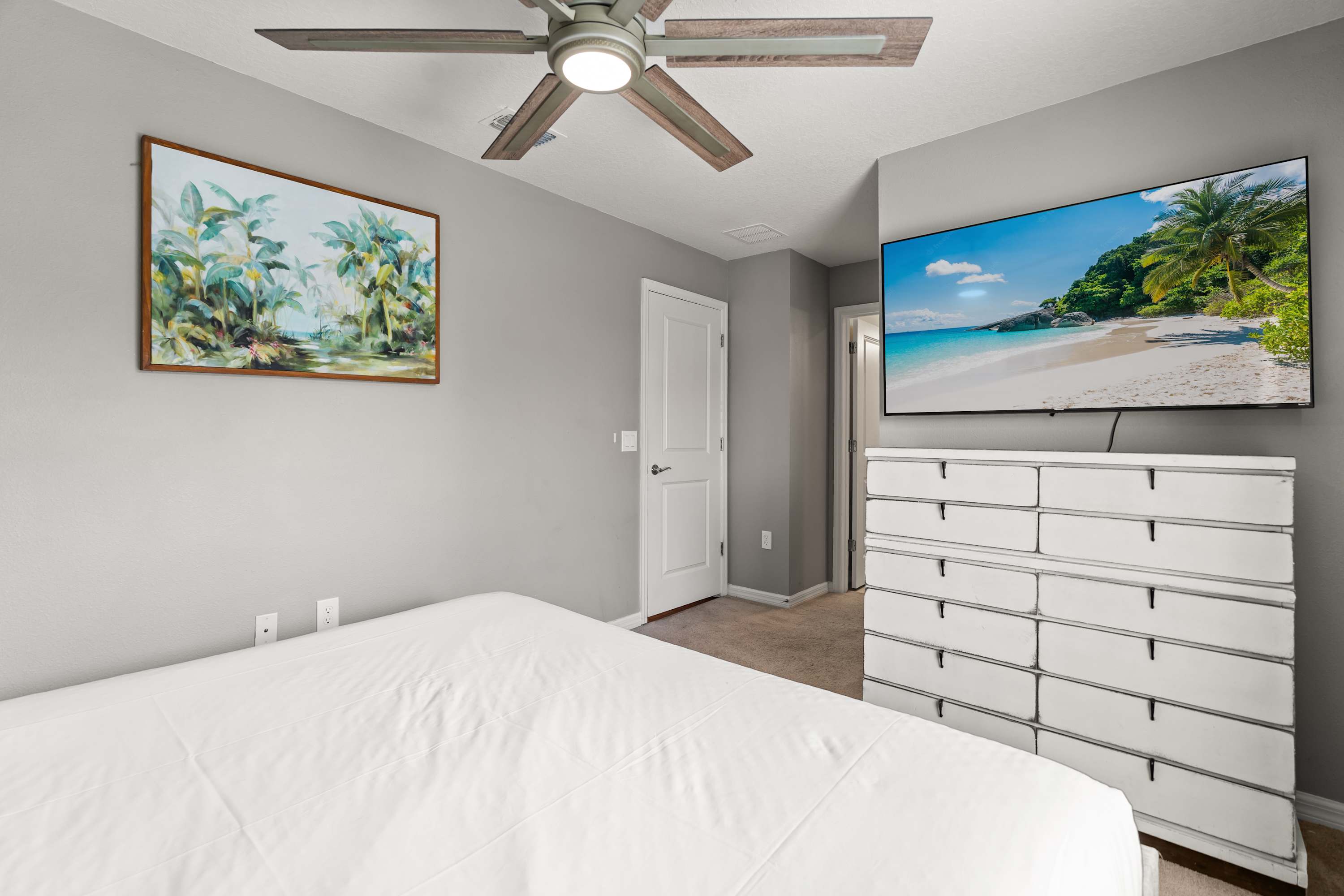 A modern bedroom with light gray walls, featuring a bed with crisp white bedding and a white dresser with multiple storage bins. A large wall-mounted TV and tropical artwork adorn the walls, with a ceiling fan above.