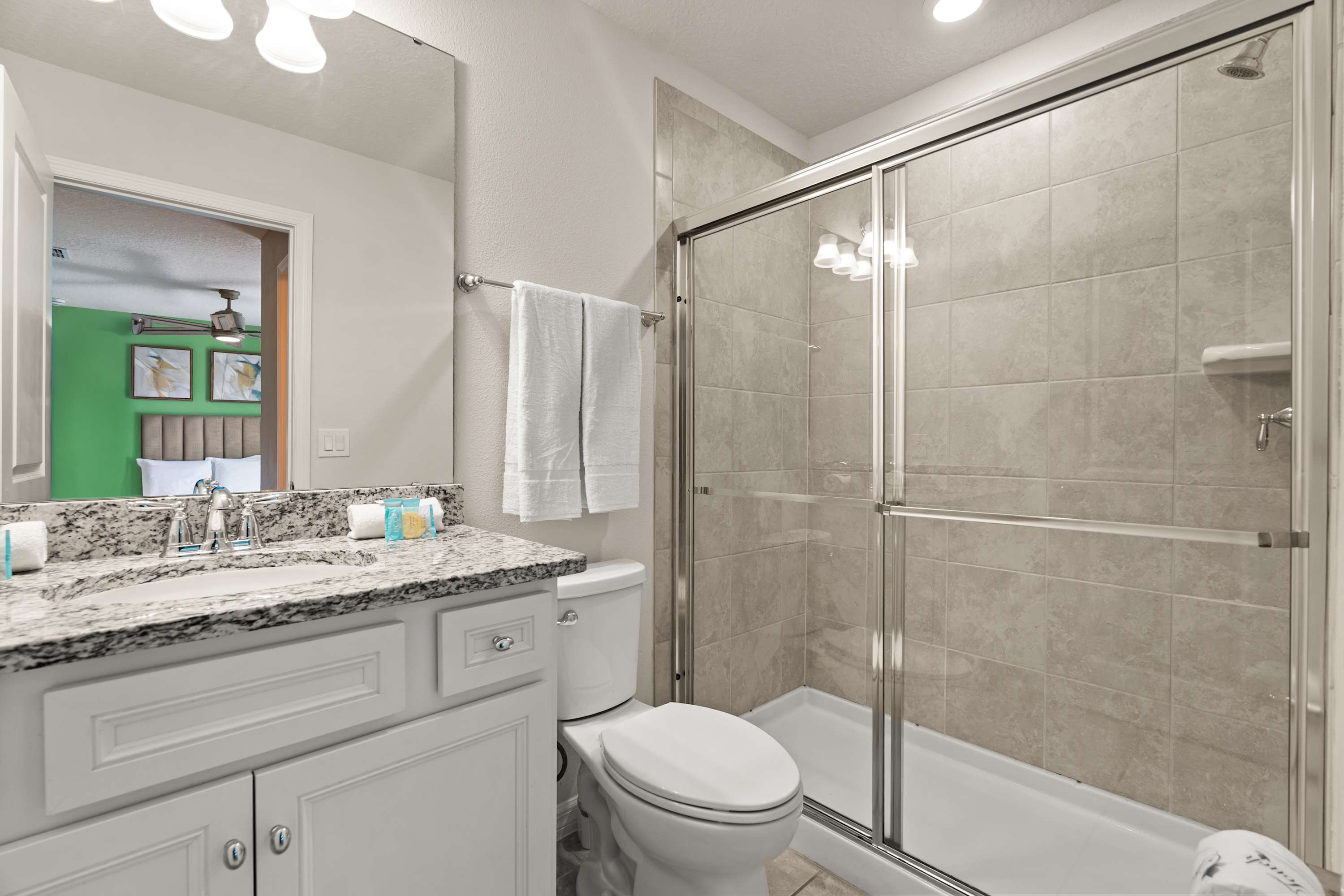 A clean bathroom with a white vanity, speckled granite countertop, and a single sink. A glass-enclosed shower with light gray tiles and a toilet are visible, reflecting a green-walled bedroom.