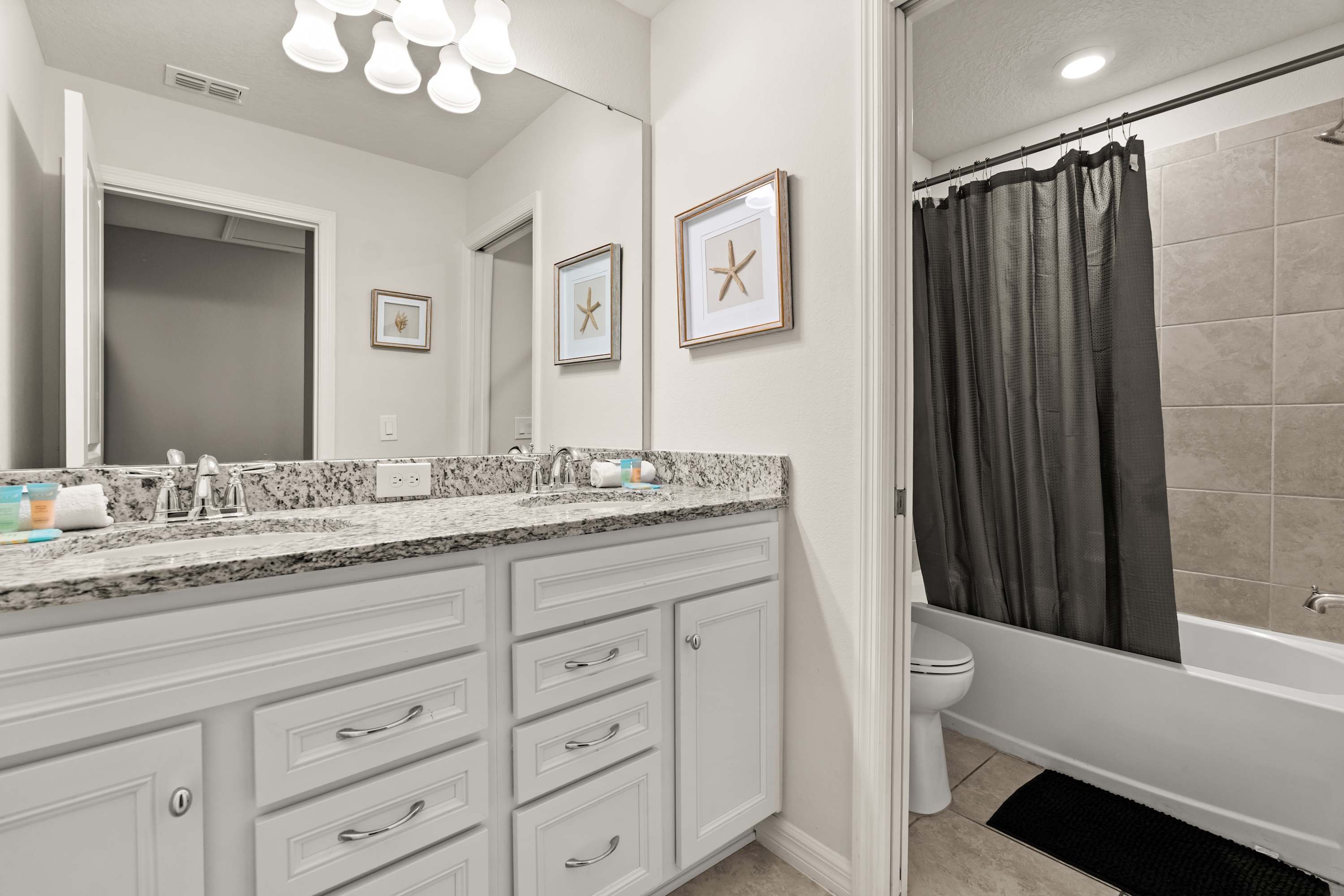 A bright bathroom features a double vanity with a granite countertop, white shaker-style cabinets, and a large mirror, with a separate area containing a toilet and a shower/tub combo visible through an open doorway.