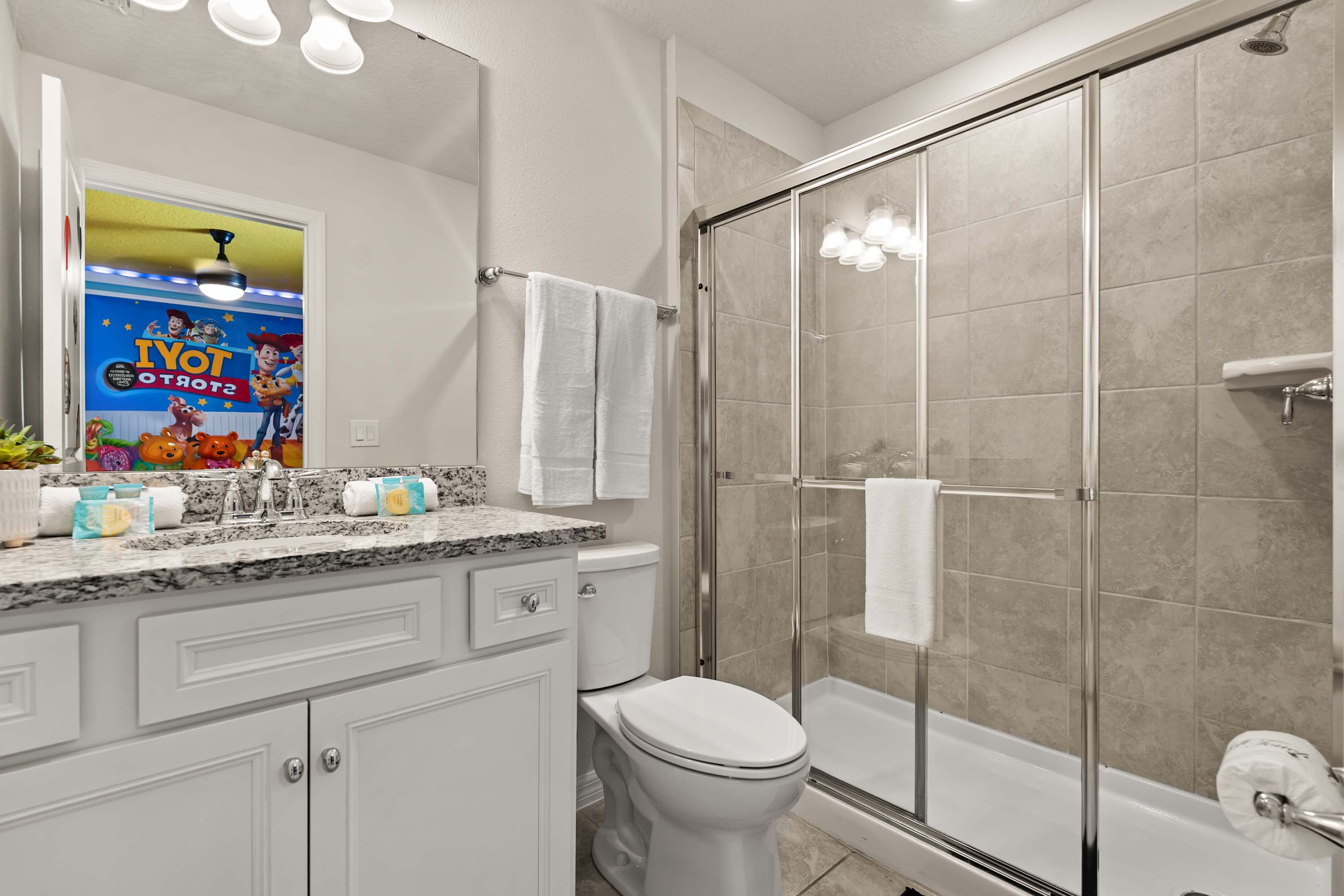 A bright bathroom featuring a white vanity with a speckled granite countertop, a single sink, and a large mirror. A glass-enclosed shower with light gray tiled walls and a toilet are visible, reflecting a "Toy Story" themed bedroom.