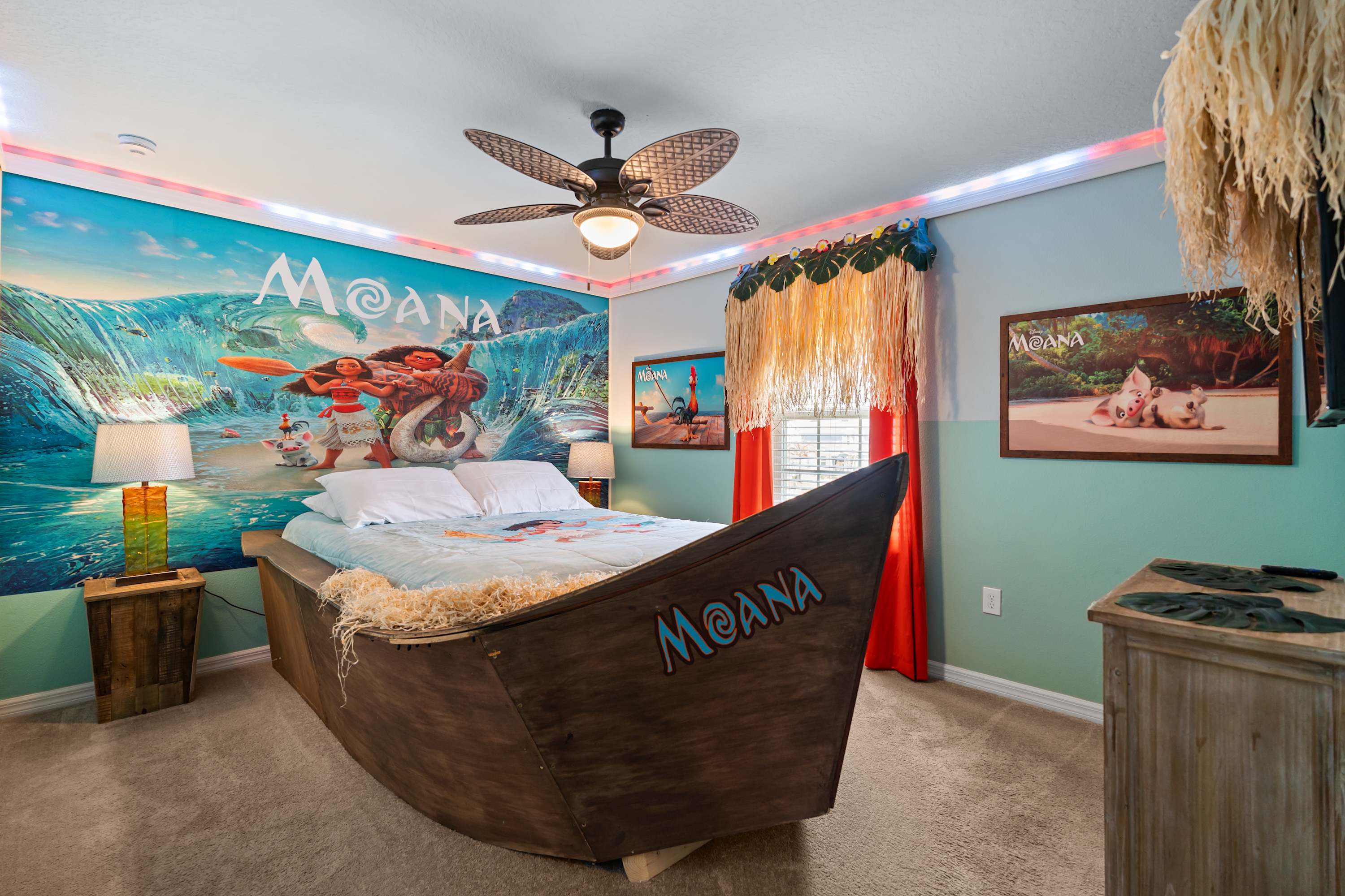 A vibrant Moana-themed bedroom features a unique boat-shaped bed with a light blue comforter, a large mural of Moana and characters on the wall, and a tropical ceiling fan with woven blades.