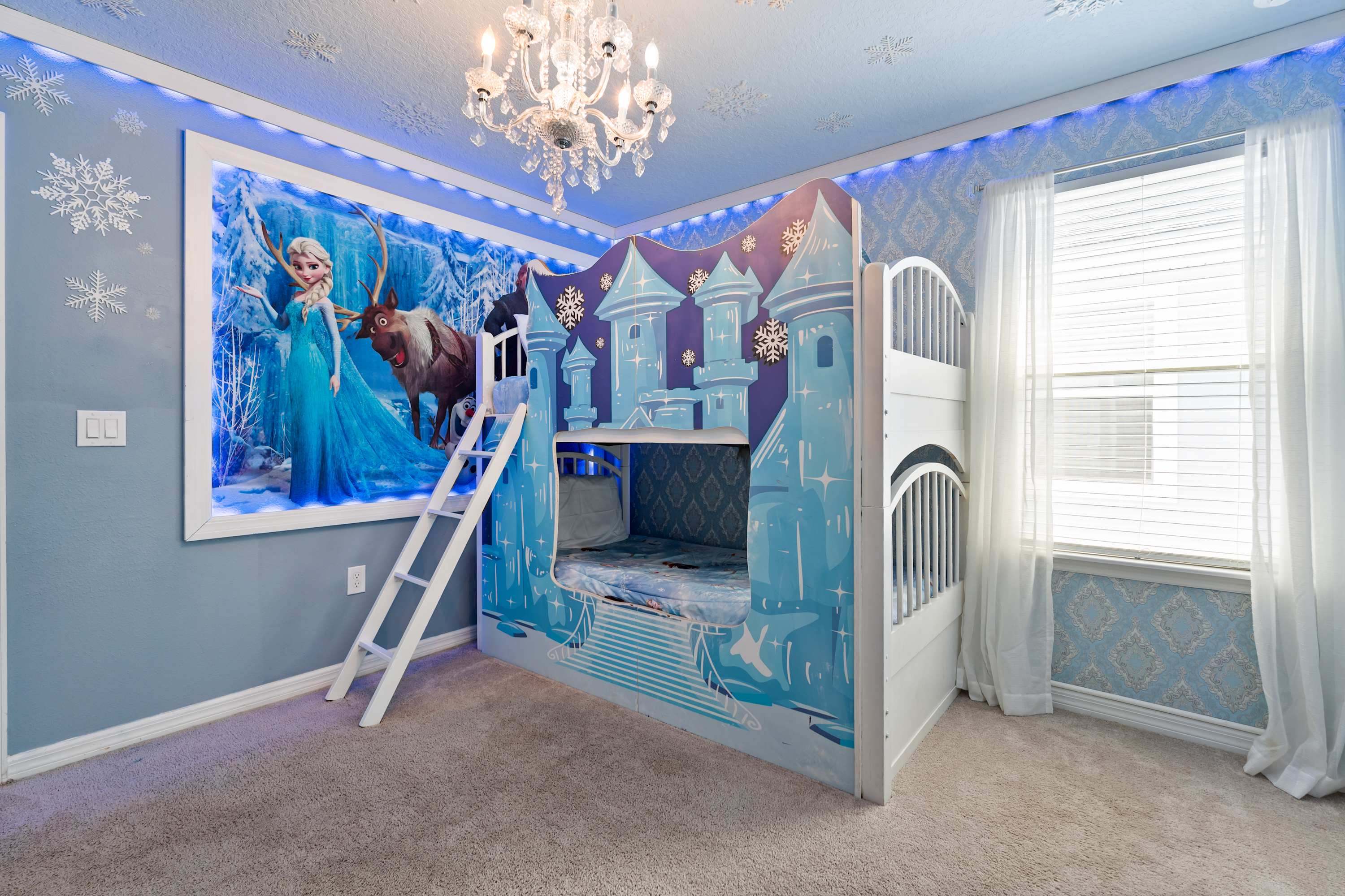 A Frozen-themed bedroom features a castle-shaped bunk bed, a large illuminated mural of Elsa and Sven, a crystal chandelier, and snowflake decals on the light blue walls and ceiling.