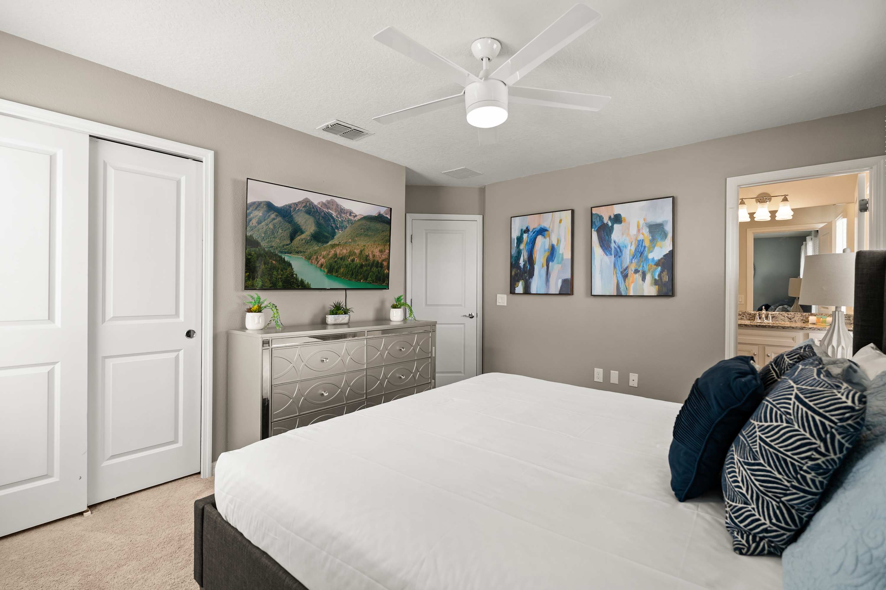 A view of the bedroom showing a large TV mounted above a mirrored dresser, with a dark upholstered bed in the foreground. Two white closet doors are visible on the left, and a door leads to an ensuite bathroom.