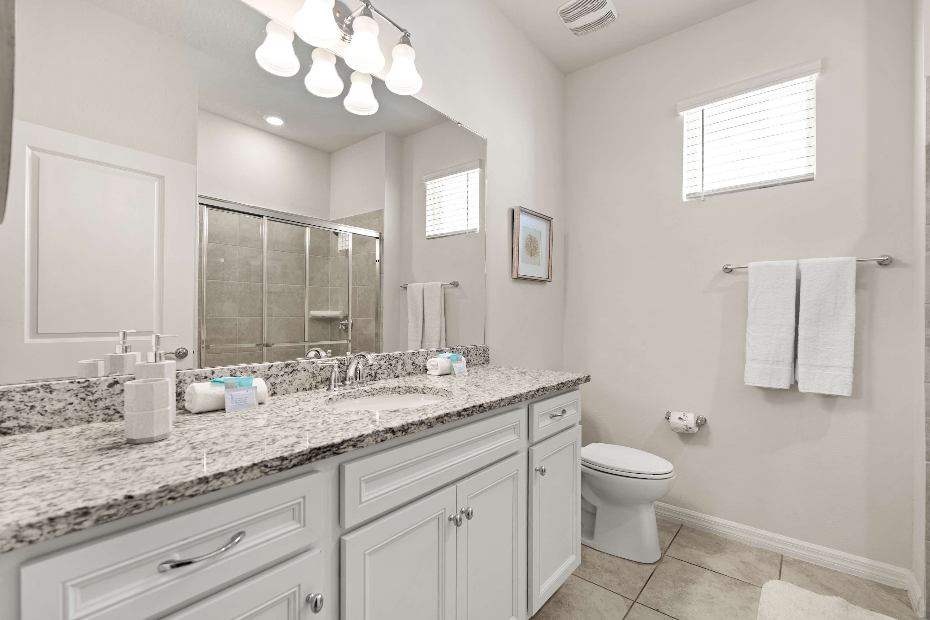 A modern bathroom features a white vanity with a granite countertop and a single sink, a large mirror, a toilet, and a glass-enclosed shower, all under bright lighting.