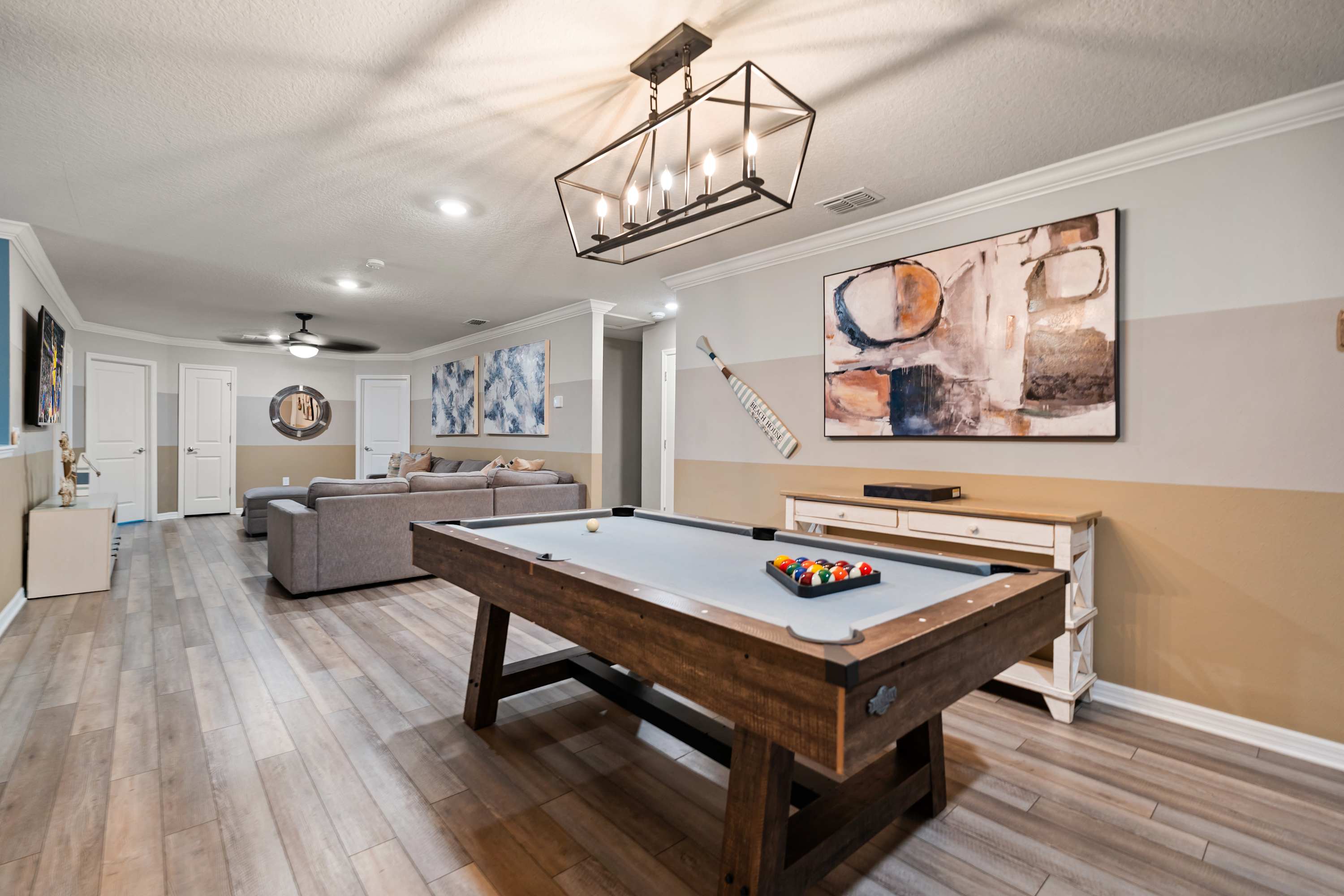 A spacious game room featuring a wooden pool table with a light blue felt top, illuminated by a modern black chandelier. A cozy living area with a gray sectional sofa is visible in the background, all set on light wood-look flooring.