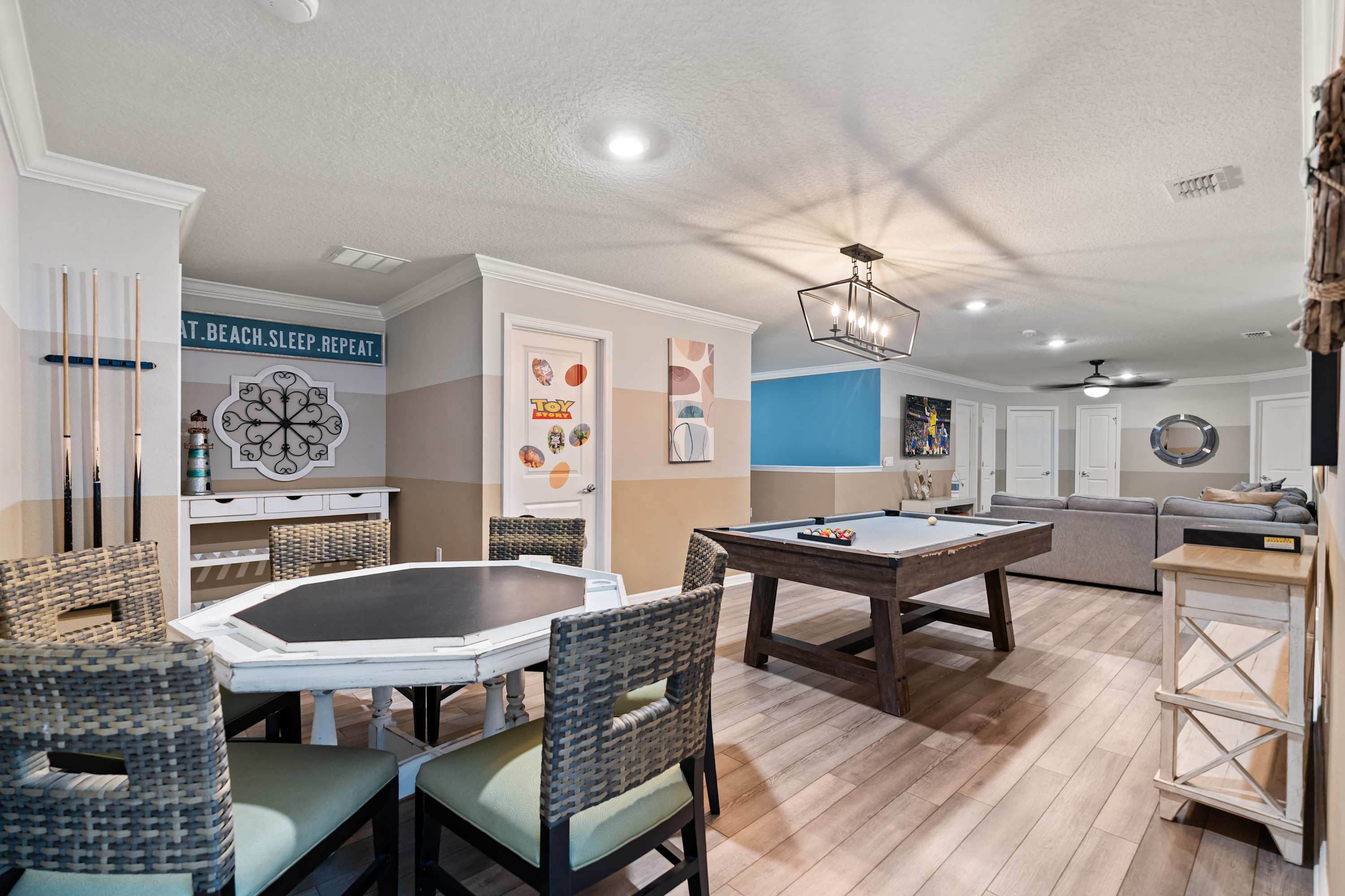Spacious game room featuring a wooden pool table, a hexagonal card table with wicker chairs, and light wood-look flooring, with a seating area visible in the background.