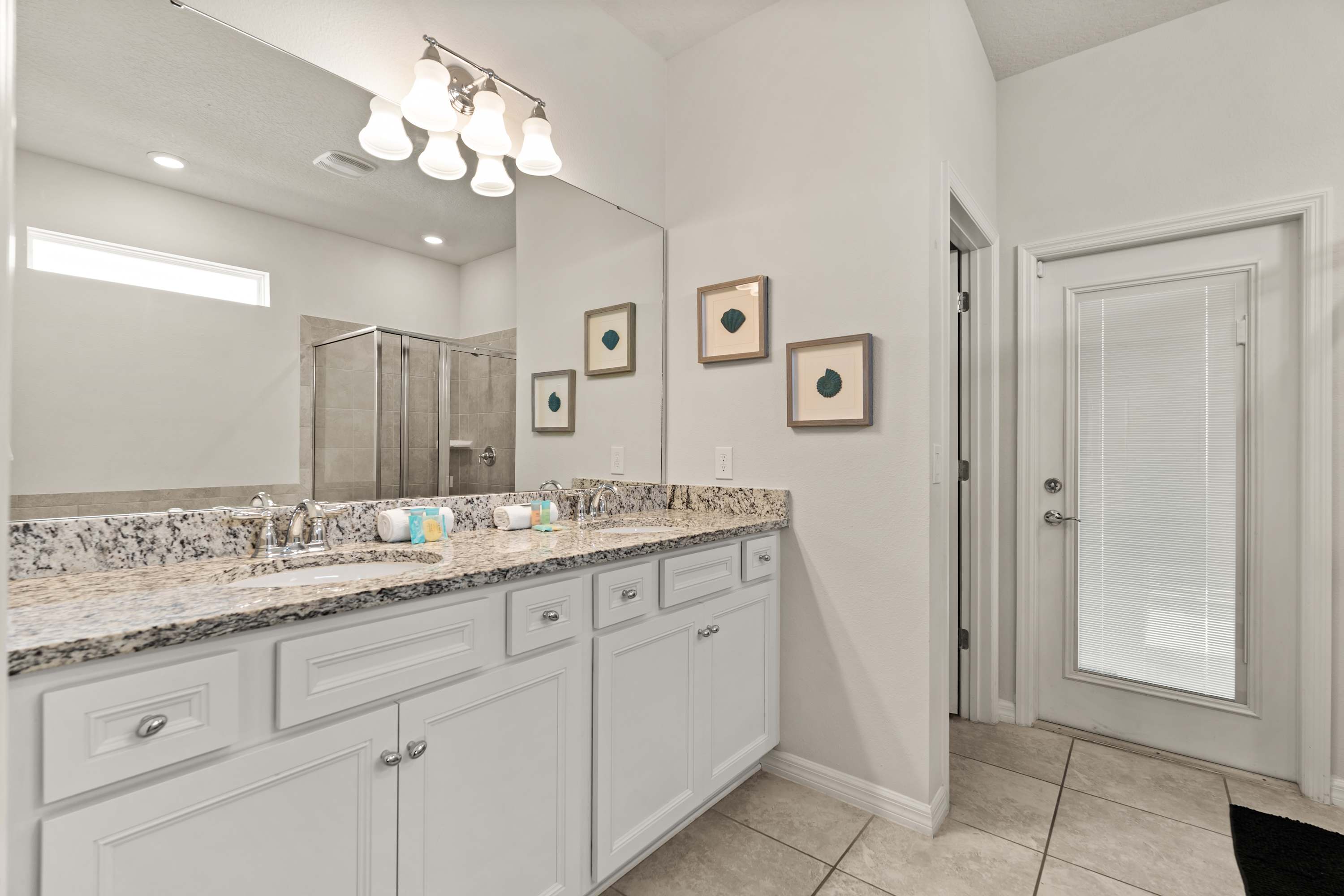 A bright bathroom with a double vanity featuring a granite countertop and white cabinets, under a large mirror with a multi-bulb light fixture. The glass shower and soaking tub are reflected in the mirror.