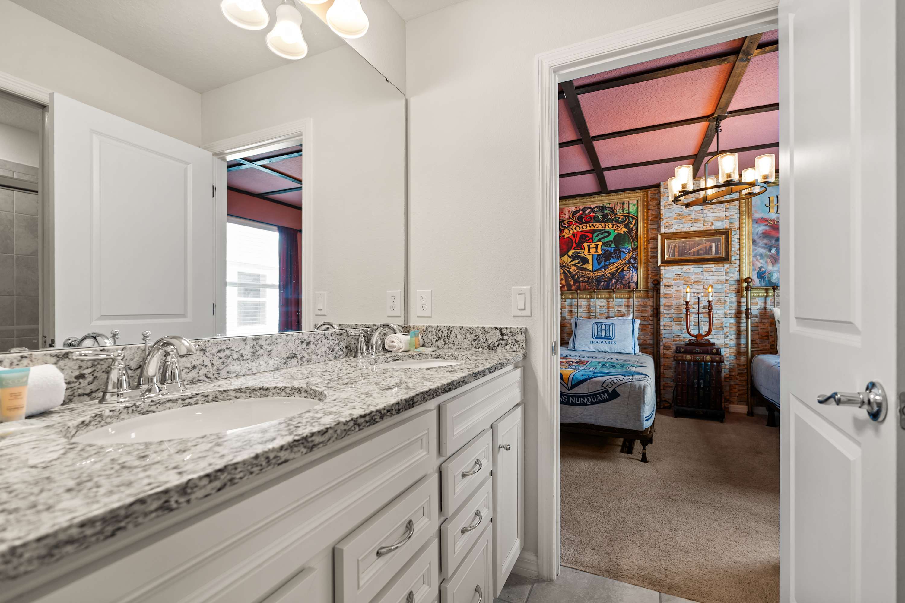 A bright bathroom features a double vanity with a granite countertop and two sinks, white shaker-style cabinets, and a large mirror, with a Harry Potter-themed bedroom visible through an open doorway.