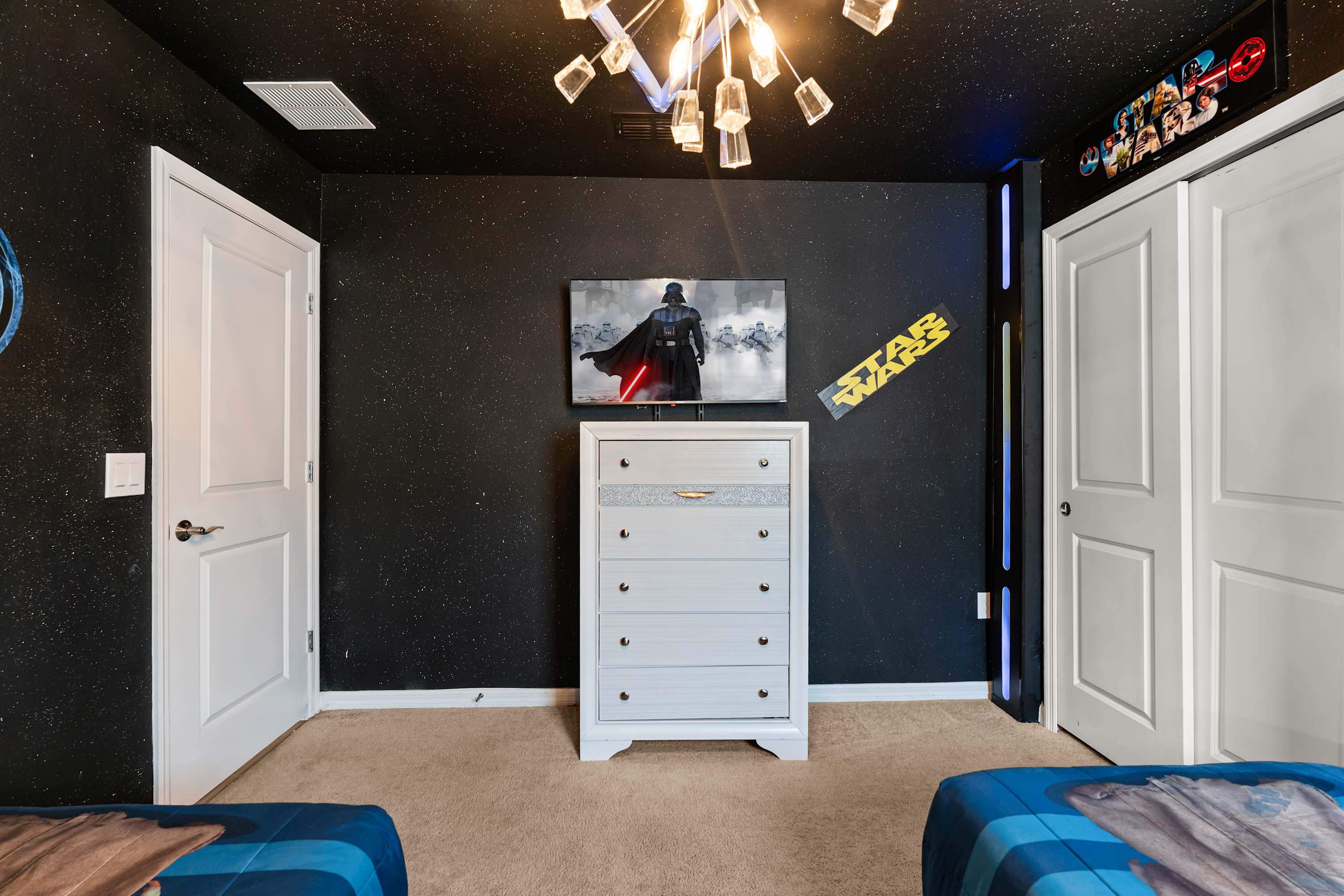 This view of the Star Wars-themed bedroom shows a white dresser with a wall-mounted TV displaying Darth Vader, a dark starry ceiling, and white closet doors, with blue LED strip lighting accents.