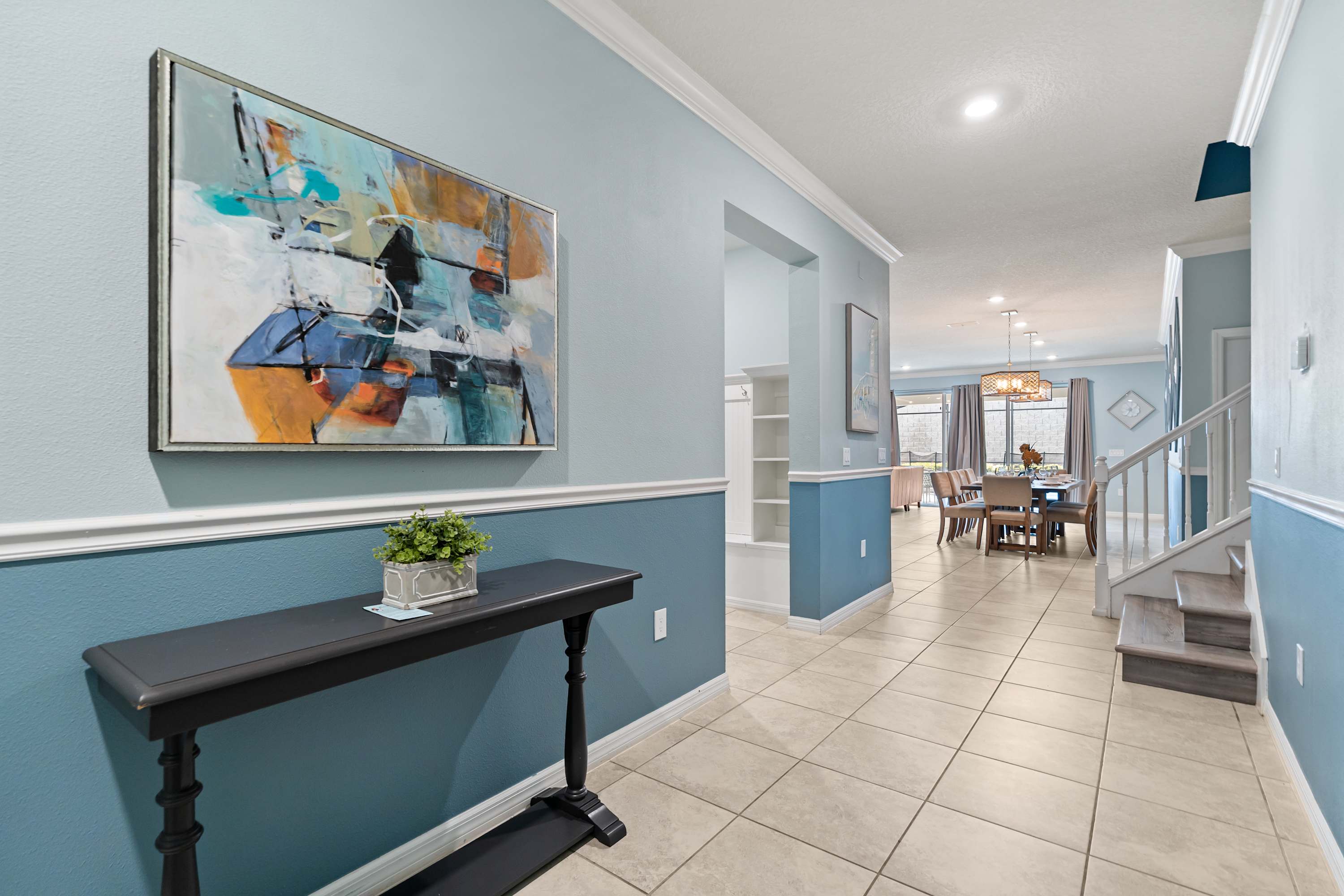 Inviting foyer with light blue and darker blue two-tone walls, featuring a black console table with a small plant, a large abstract painting, and tile flooring, leading into the open-concept living and dining areas.