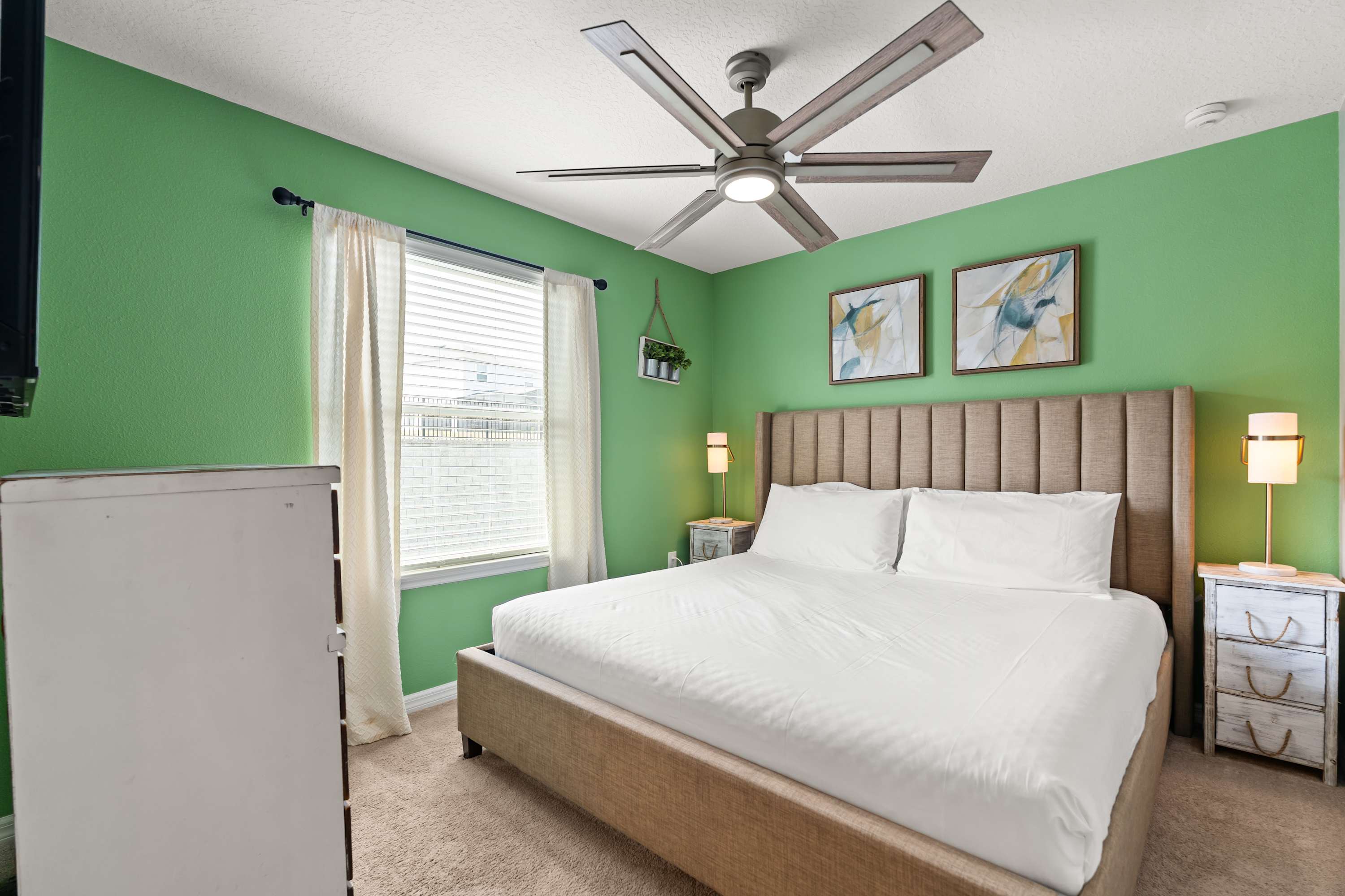 A vibrant bedroom with bright green walls, featuring a king-sized bed with a beige upholstered headboard. Two white nightstands with lamps flank the bed, and a ceiling fan hangs overhead.