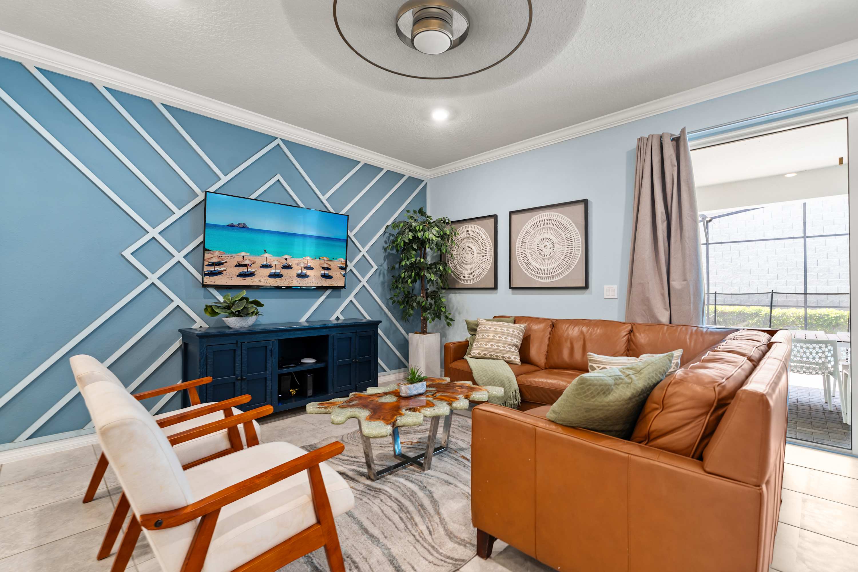 Stylish living room featuring a brown leather sectional sofa, a unique live-edge coffee table, and a large wall-mounted TV against a light blue accent wall with geometric white trim, offering a view to the outdoor patio.
