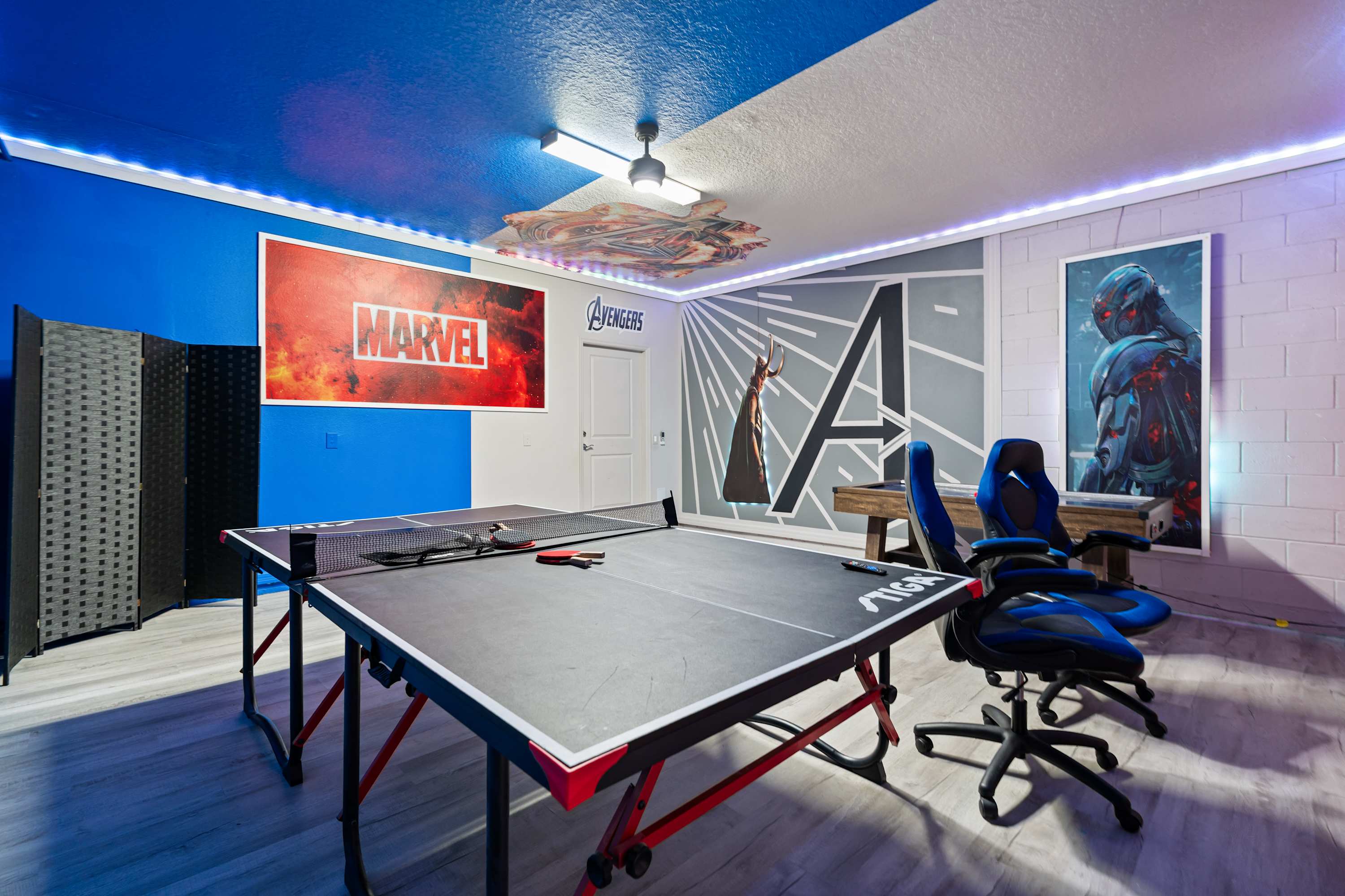 Vibrant Marvel-themed game room with a ping pong table, gaming chairs, and a large "MARVEL" sign on a blue and white wall, featuring LED strip lighting and a unique ceiling design.