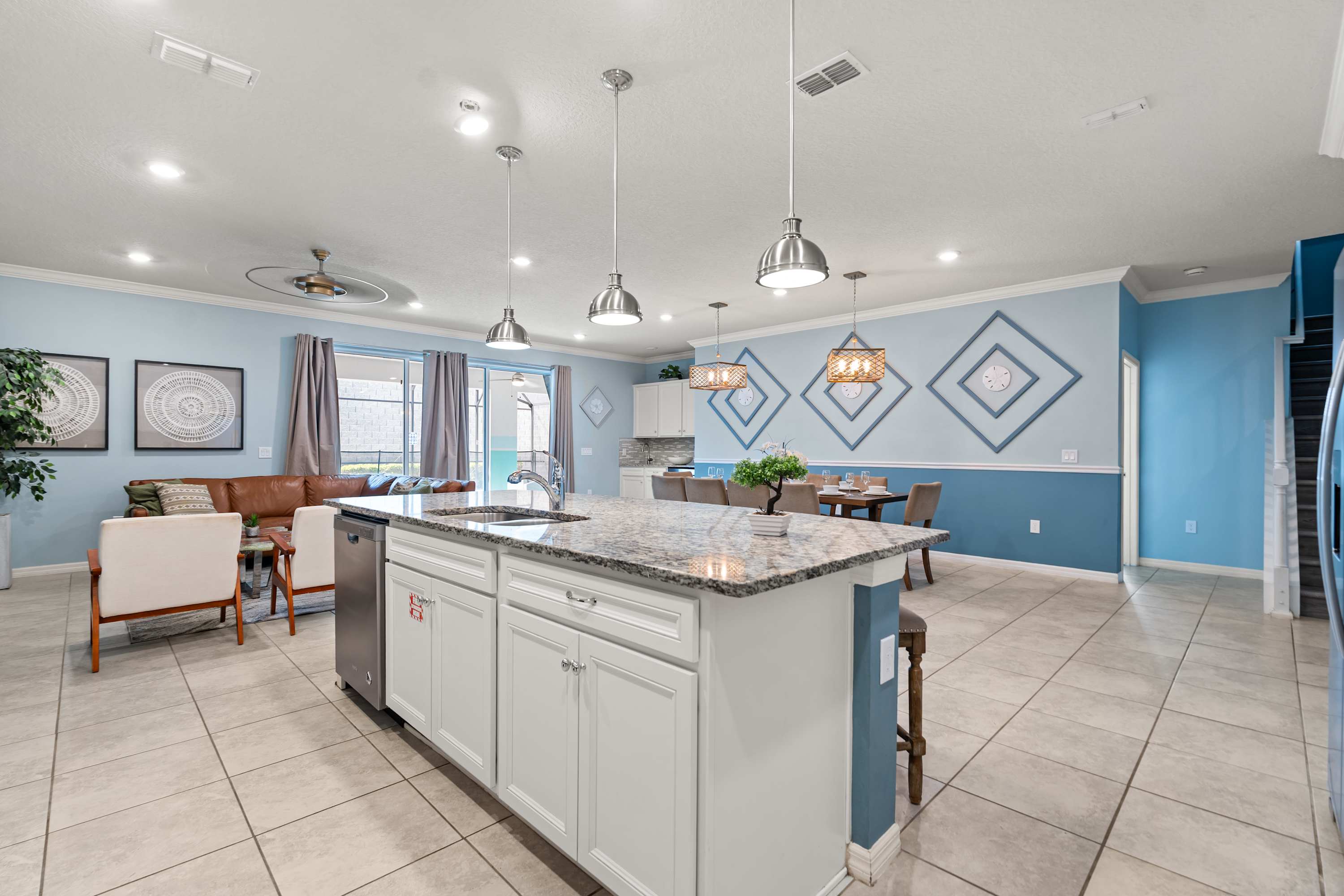 View from the kitchen island, featuring a double sink and granite countertop, looking towards a dining area with a long table and a living room with a brown leather sofa, all under recessed lighting and a ceiling fan, with sliding glass doors leading outside.