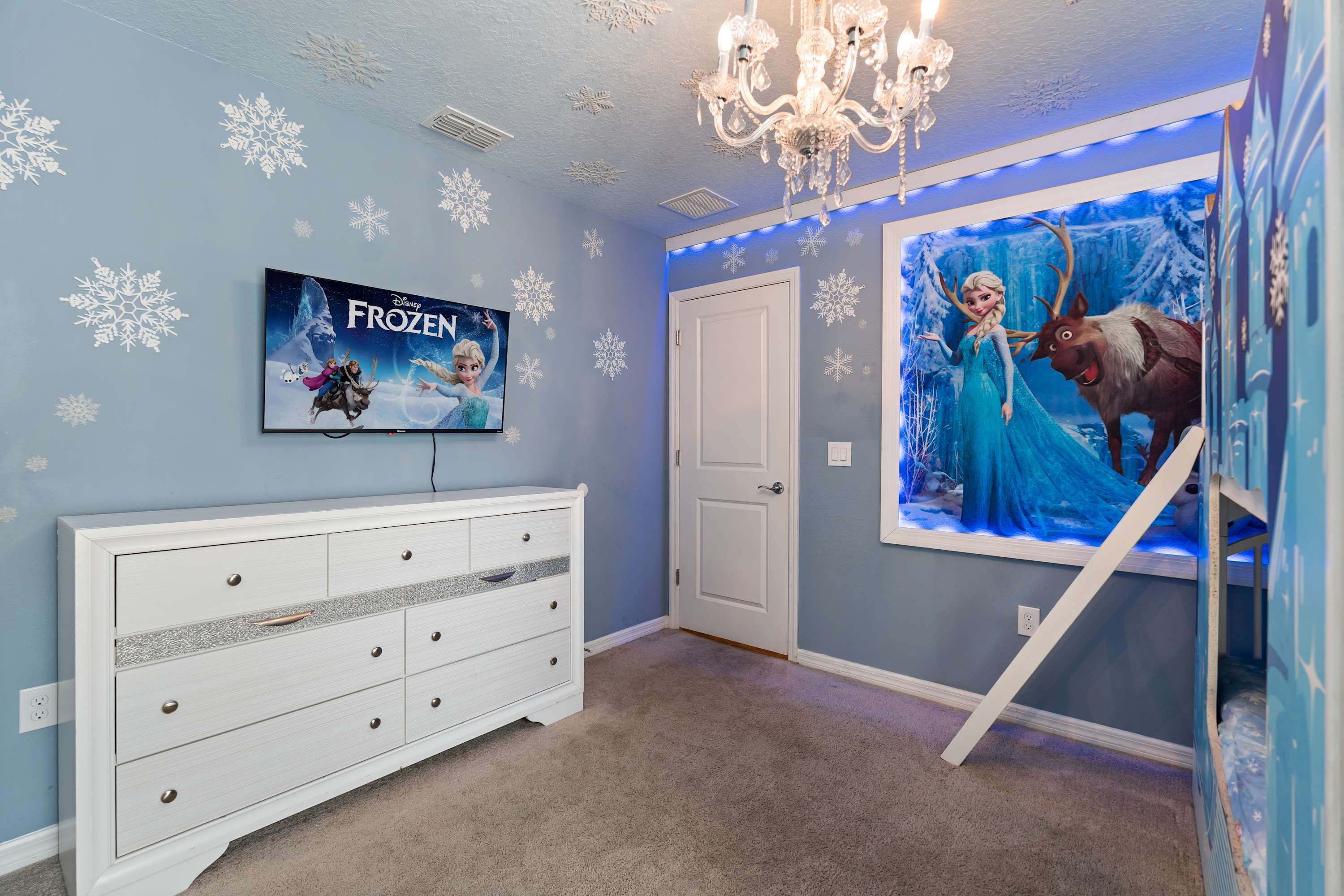 This view of the Frozen-themed bedroom highlights a white dresser with a wall-mounted TV, the illuminated Elsa and Sven mural, and the light blue walls adorned with snowflake decals and blue LED strip lighting.