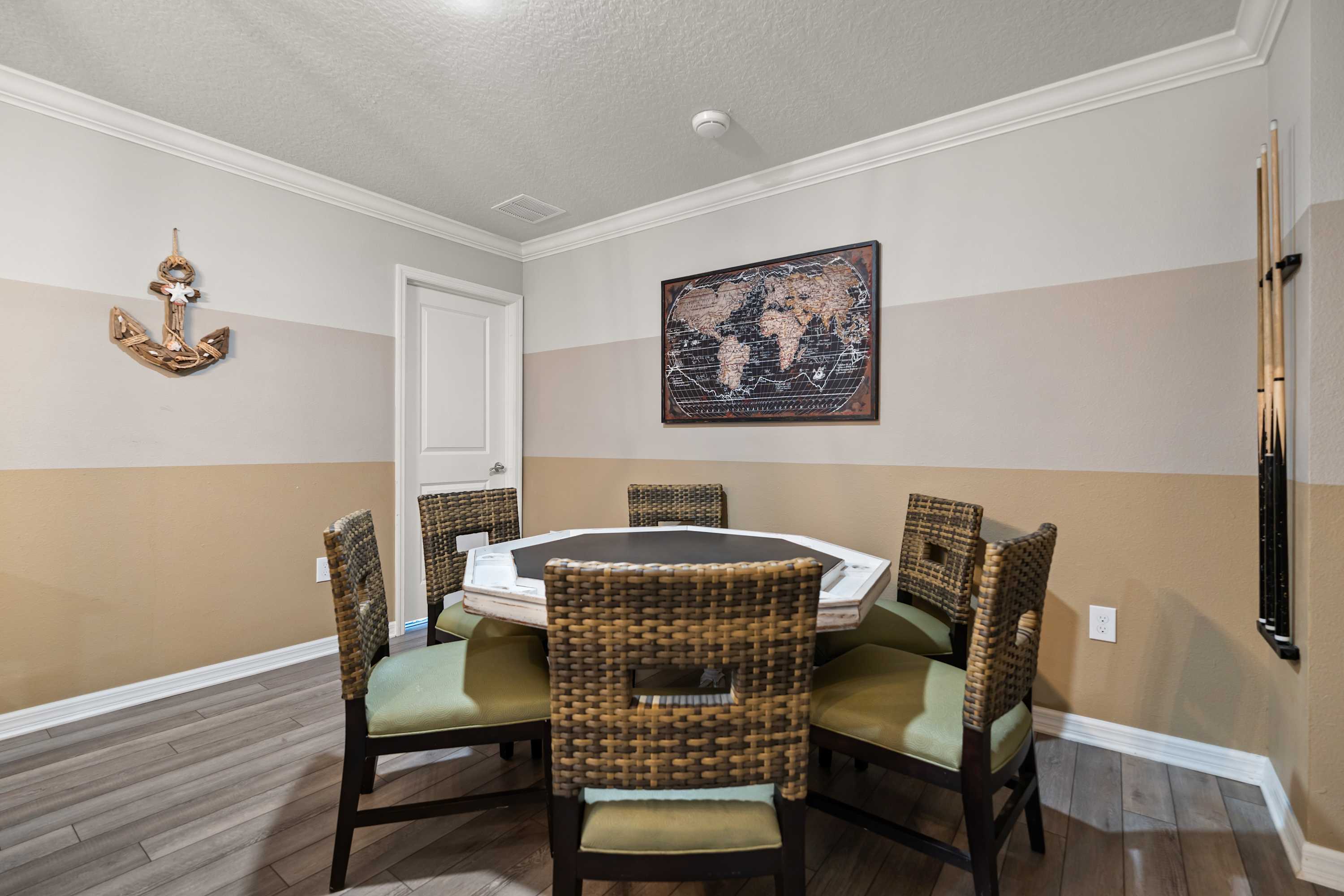 Dedicated game area with an octagonal card table and six wicker chairs, set against two-tone beige and light gray walls, featuring a world map artwork and light wood-look flooring.
