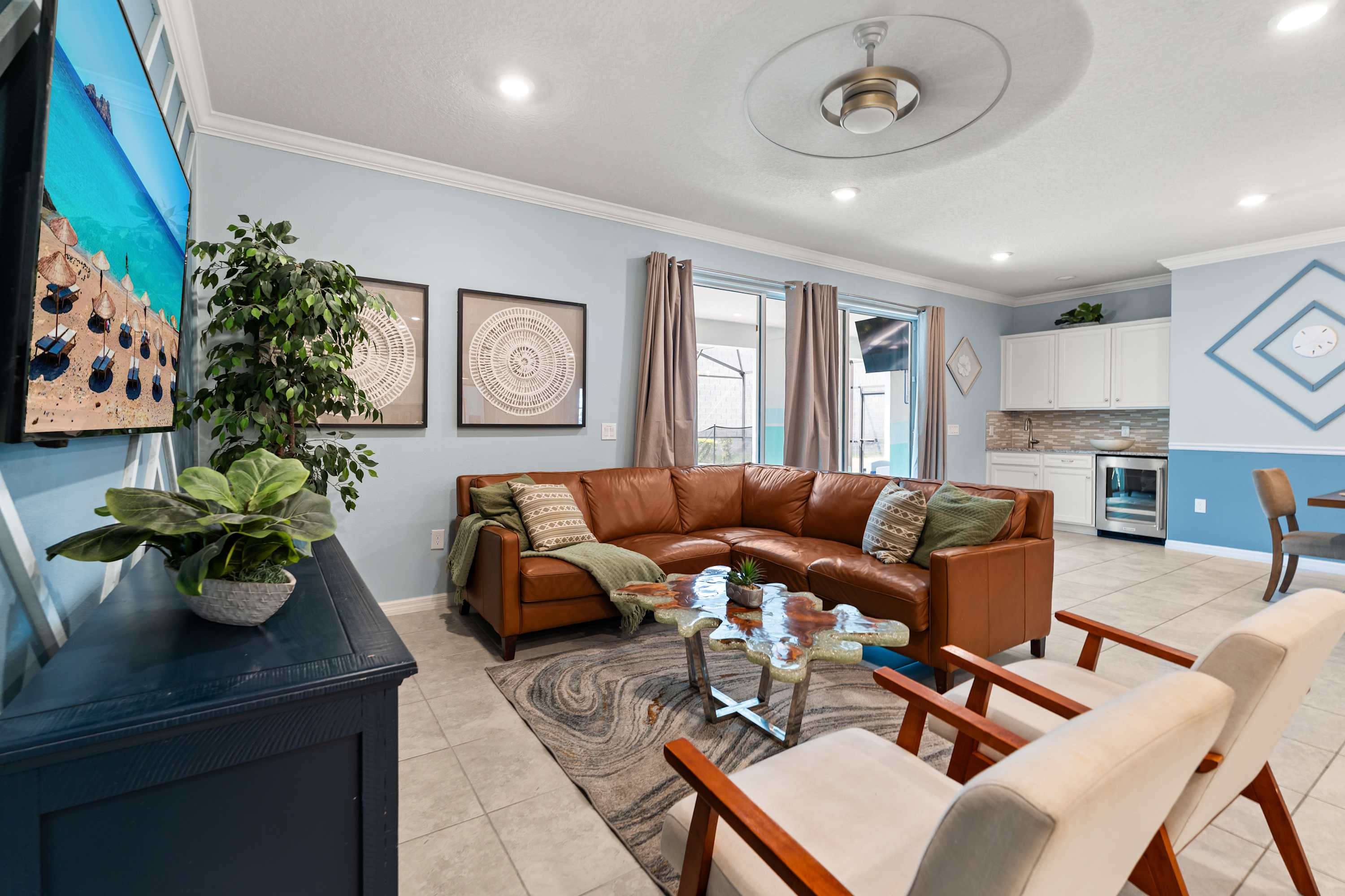 Cozy living room with a large brown leather sectional sofa, a unique live-edge coffee table, and a wall-mounted TV above a blue console, featuring light blue walls and a wet bar in the background.