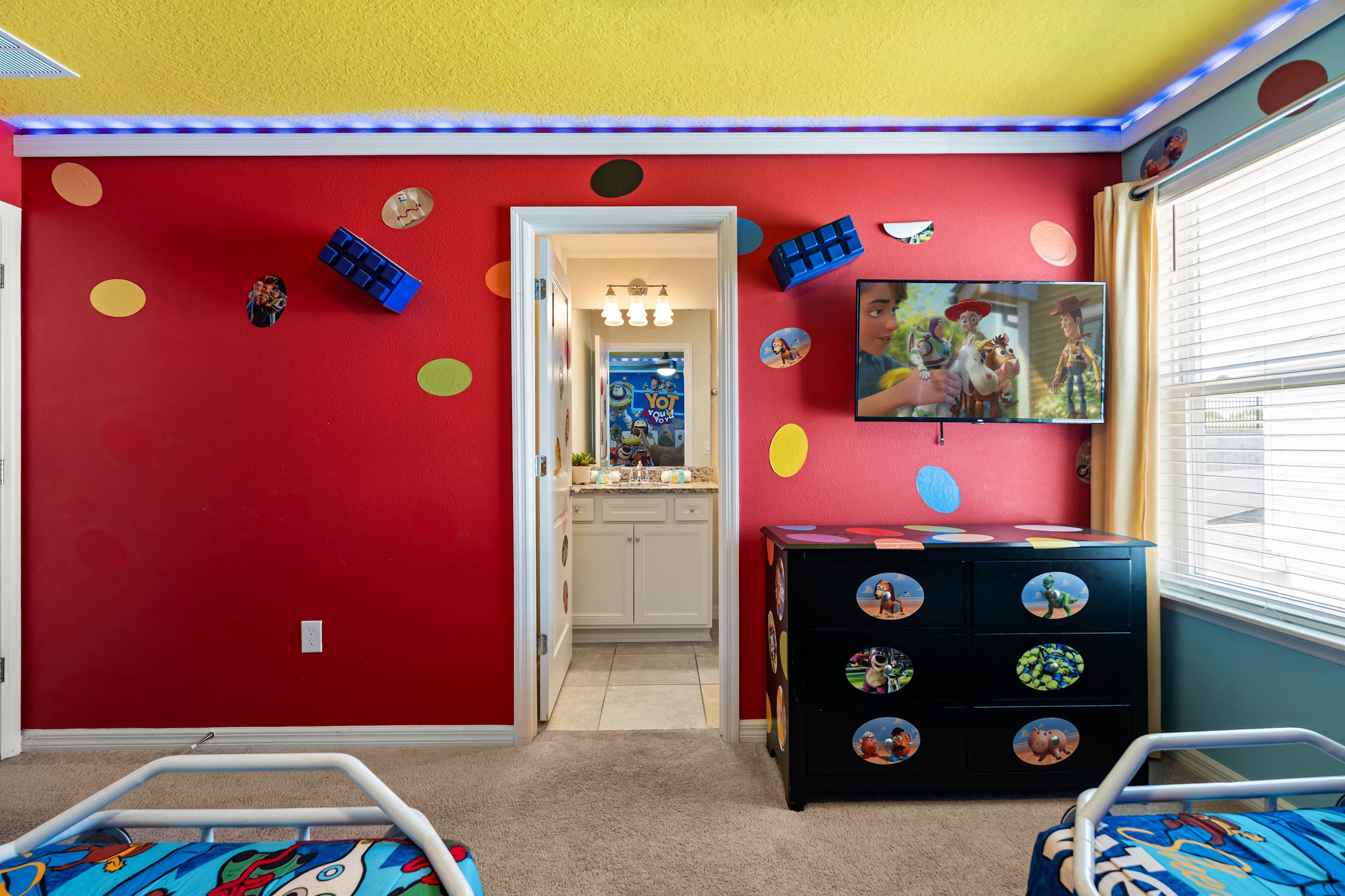 Another view of the "Toy Story" themed bedroom, showcasing a red wall with colorful polka dots and a wall-mounted TV above a black dresser. A doorway leads to an adjacent bathroom.