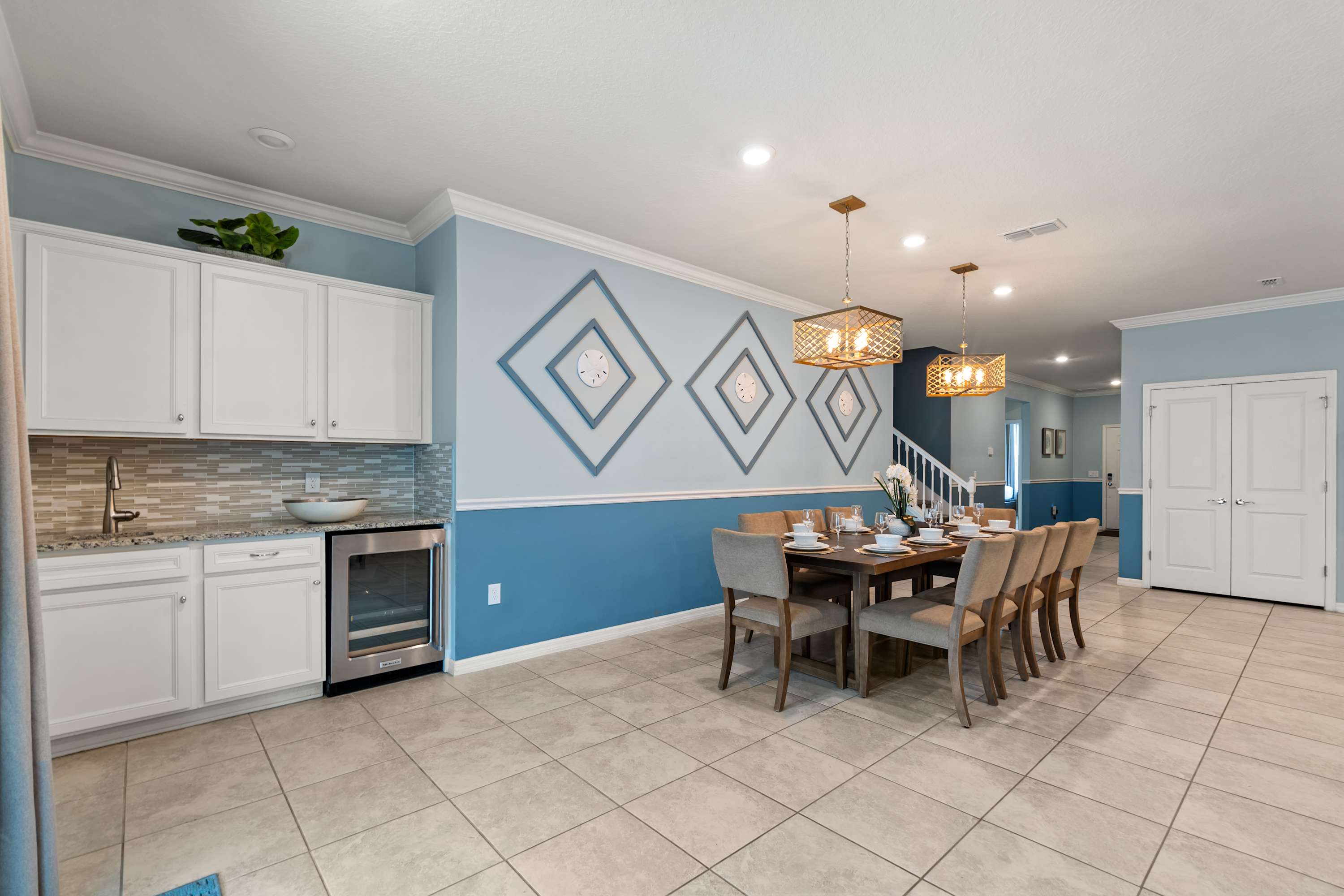 Modern dining area with a long wooden table set for eight, illuminated by two woven pendant lights, adjacent to a wet bar with white cabinets, granite countertops, and a beverage cooler, all against a blue and light blue wall with decorative diamond patterns.