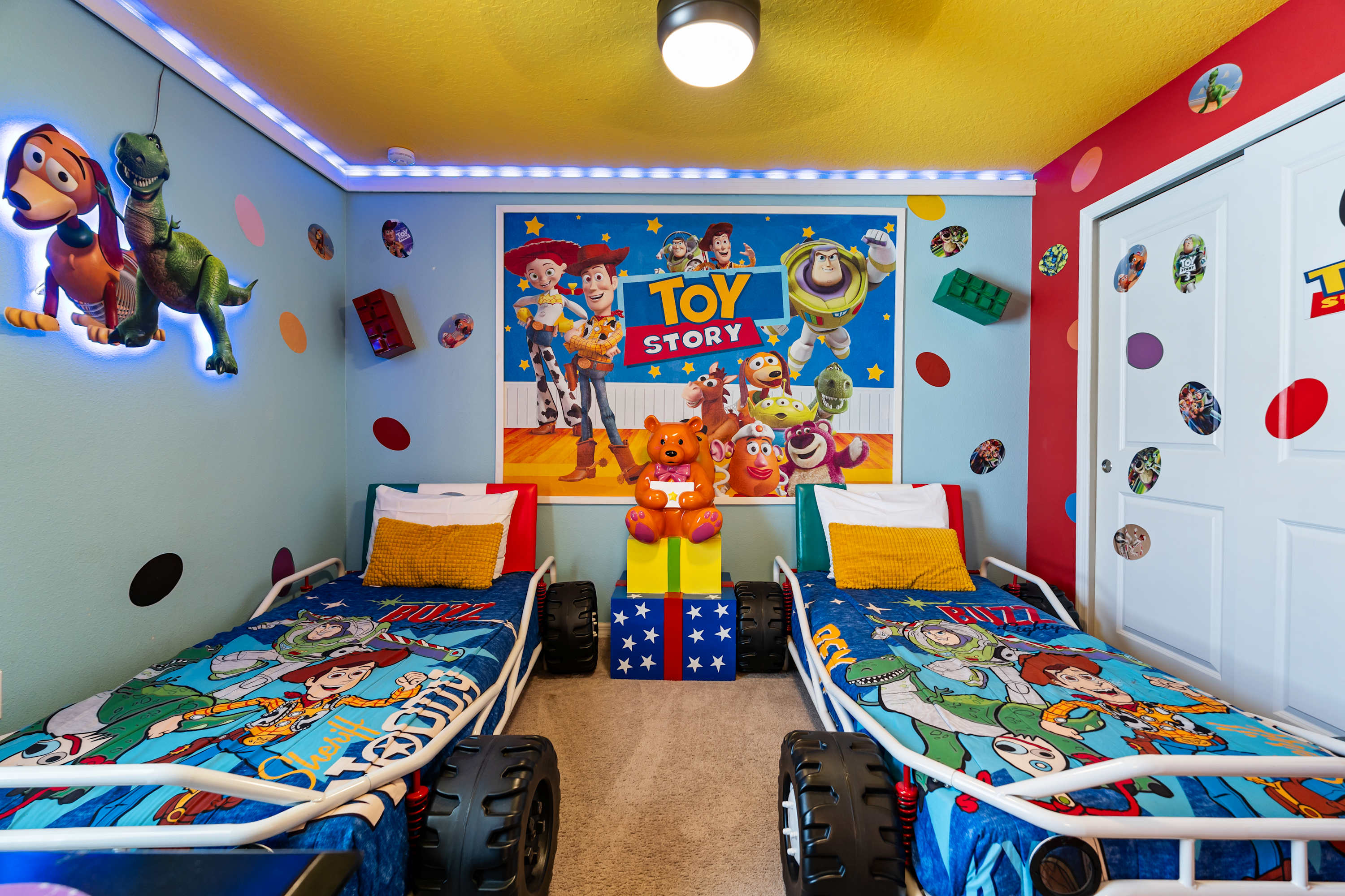 A vibrant "Toy Story" themed bedroom featuring two twin beds designed as race cars, set against blue walls with colorful polka dots and a large character mural. The room has a bright yellow ceiling and playful decorations.