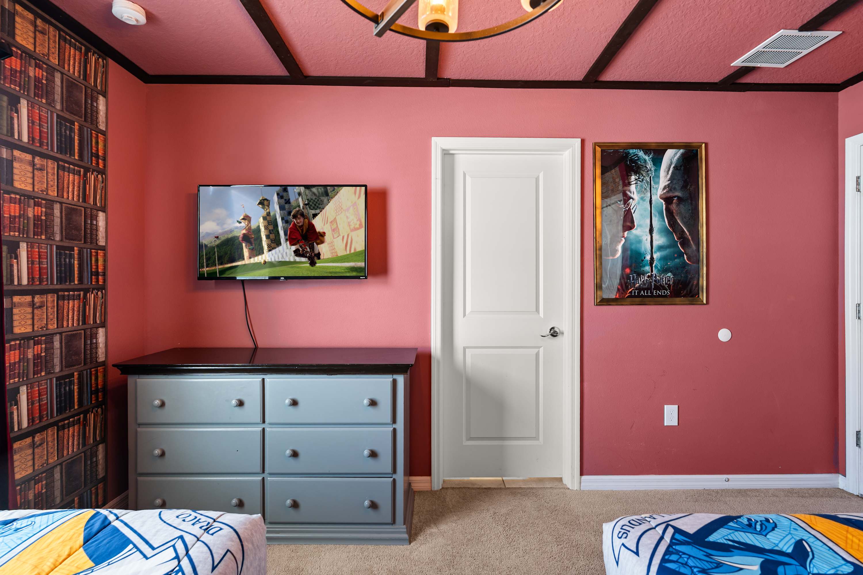 A section of the "Harry Potter" themed bedroom, showing a gray dresser with a wall-mounted TV above it. A white door is visible on the right, and bookshelf-patterned wallpaper covers the wall.