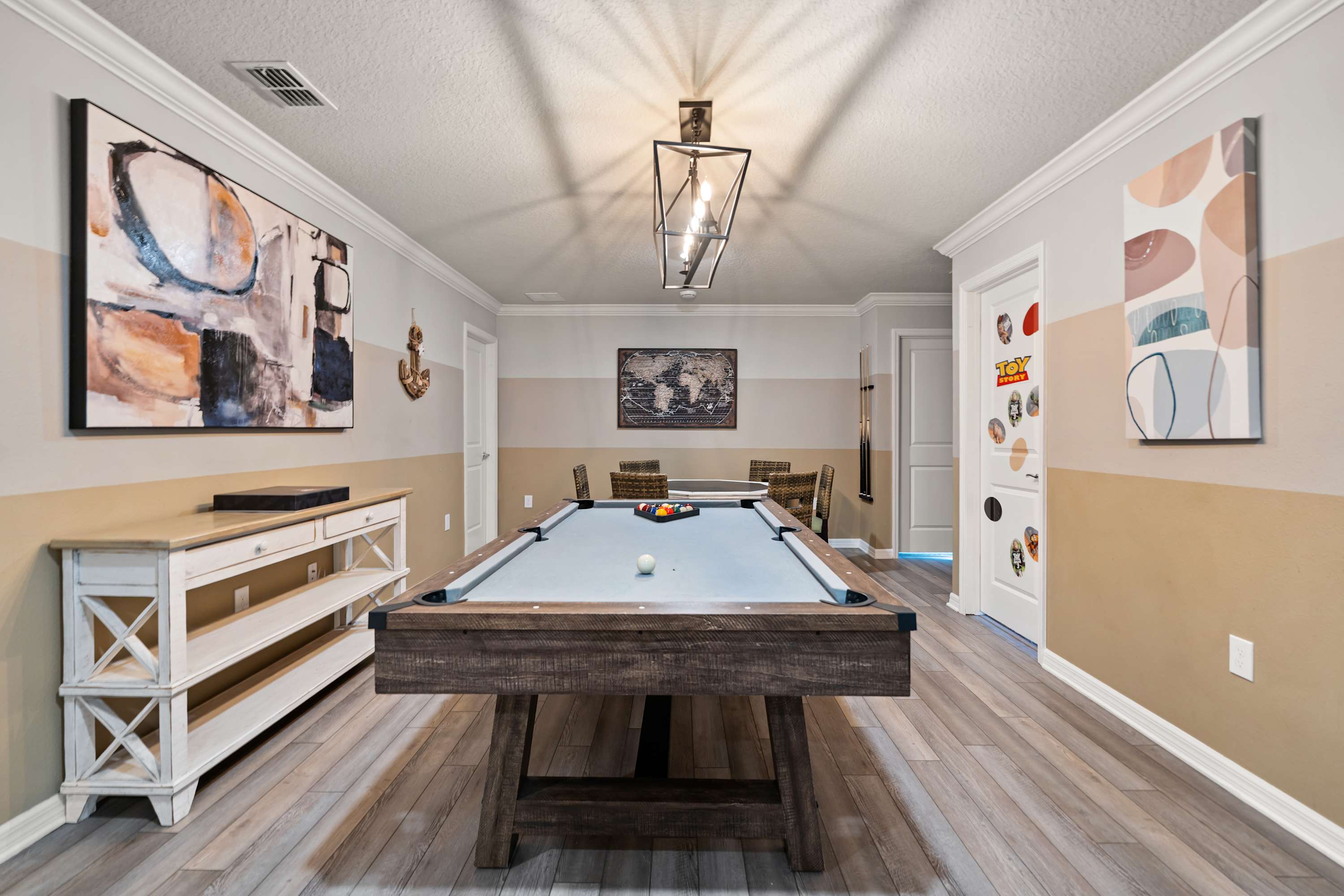 Stylish game room featuring a rustic wooden pool table with a light blue felt, illuminated by a modern linear chandelier, with two-tone walls in beige and light gray, and light wood-look flooring.