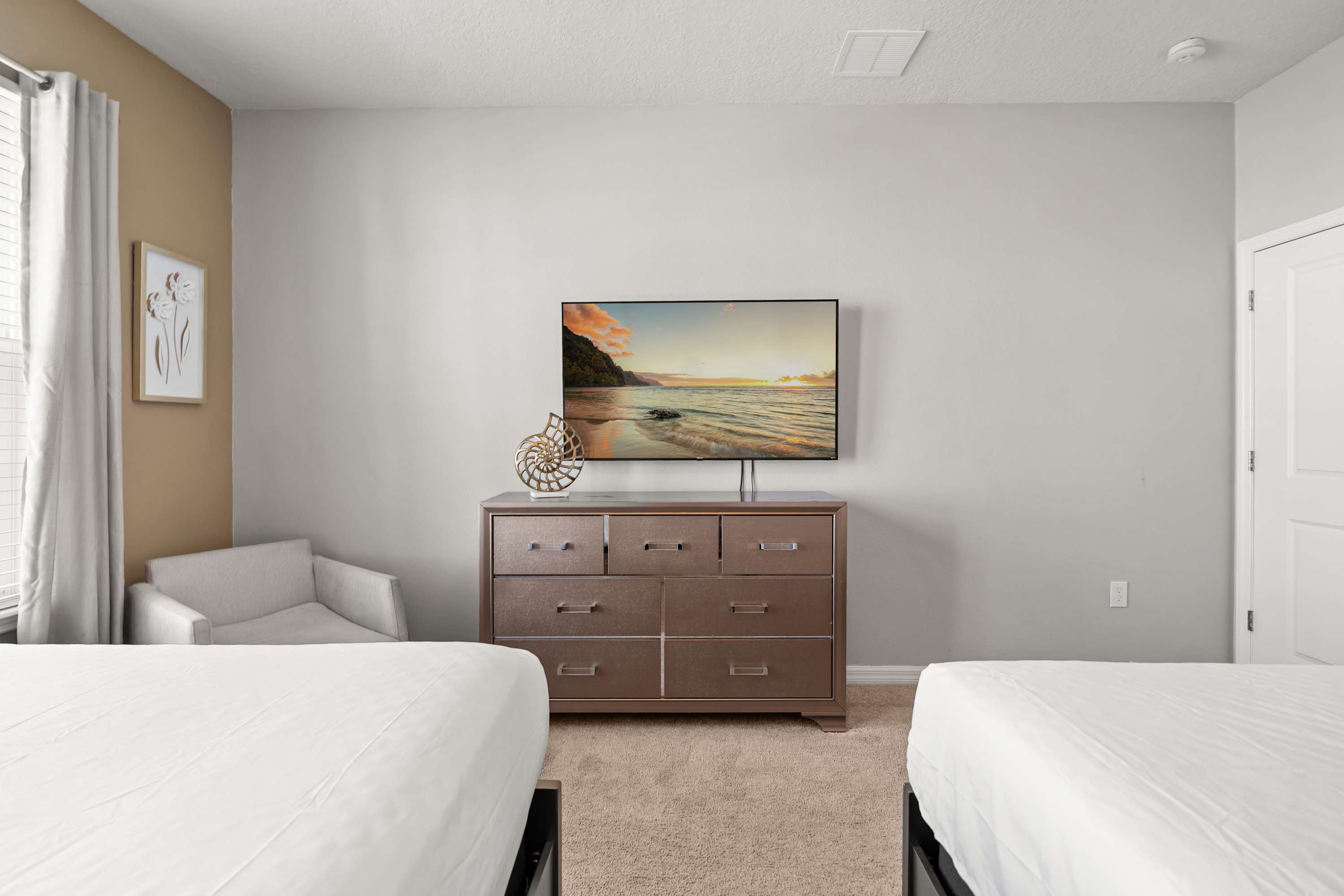 A close-up view of the bedroom shows a flat-screen TV mounted above a dark wood dresser, with a decorative shell sculpture and a light-colored armchair in the corner.