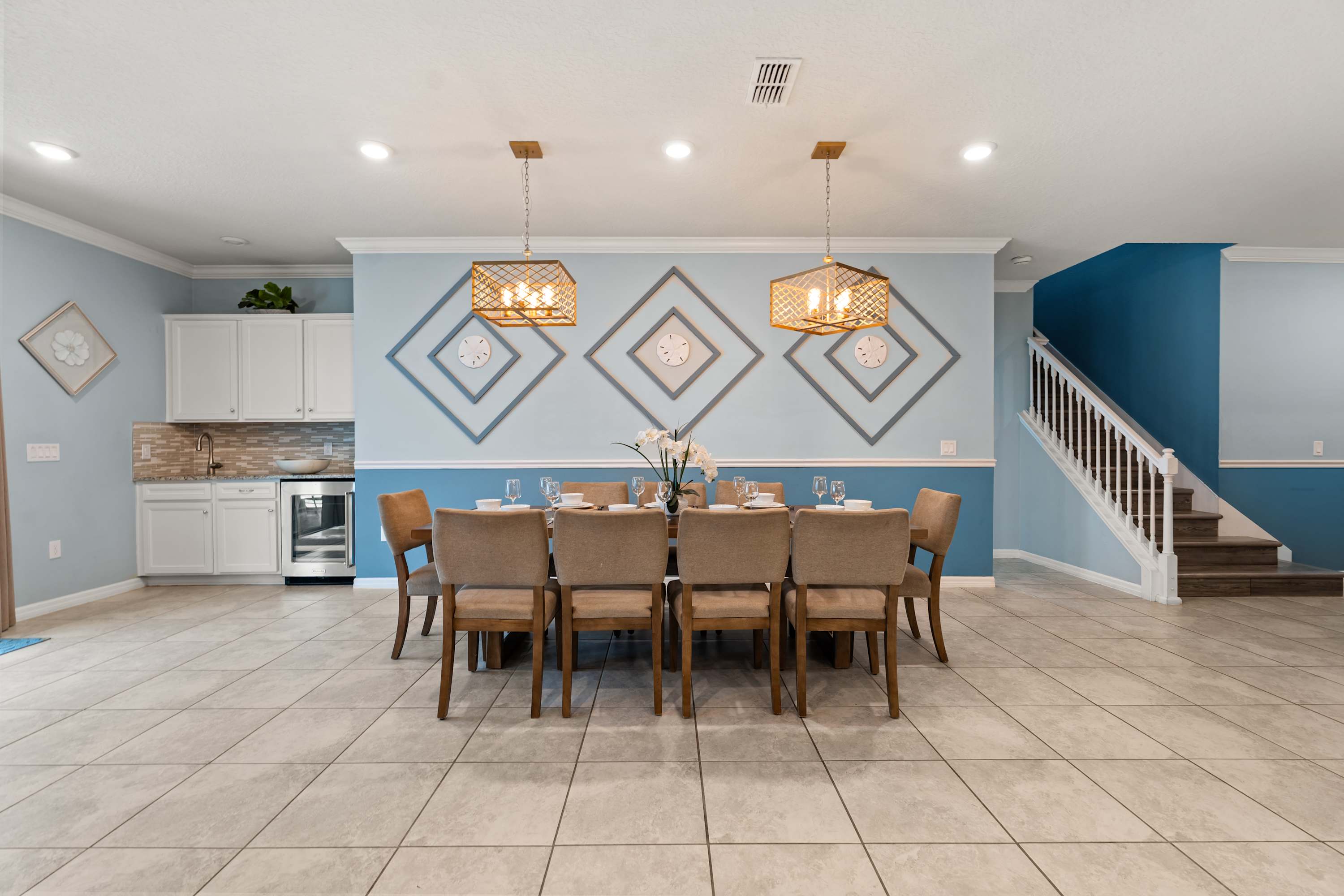 Spacious dining room featuring a long wooden dining table with seating for ten, illuminated by two modern pendant lights, set against light blue walls with geometric square decor and a wet bar in the background.