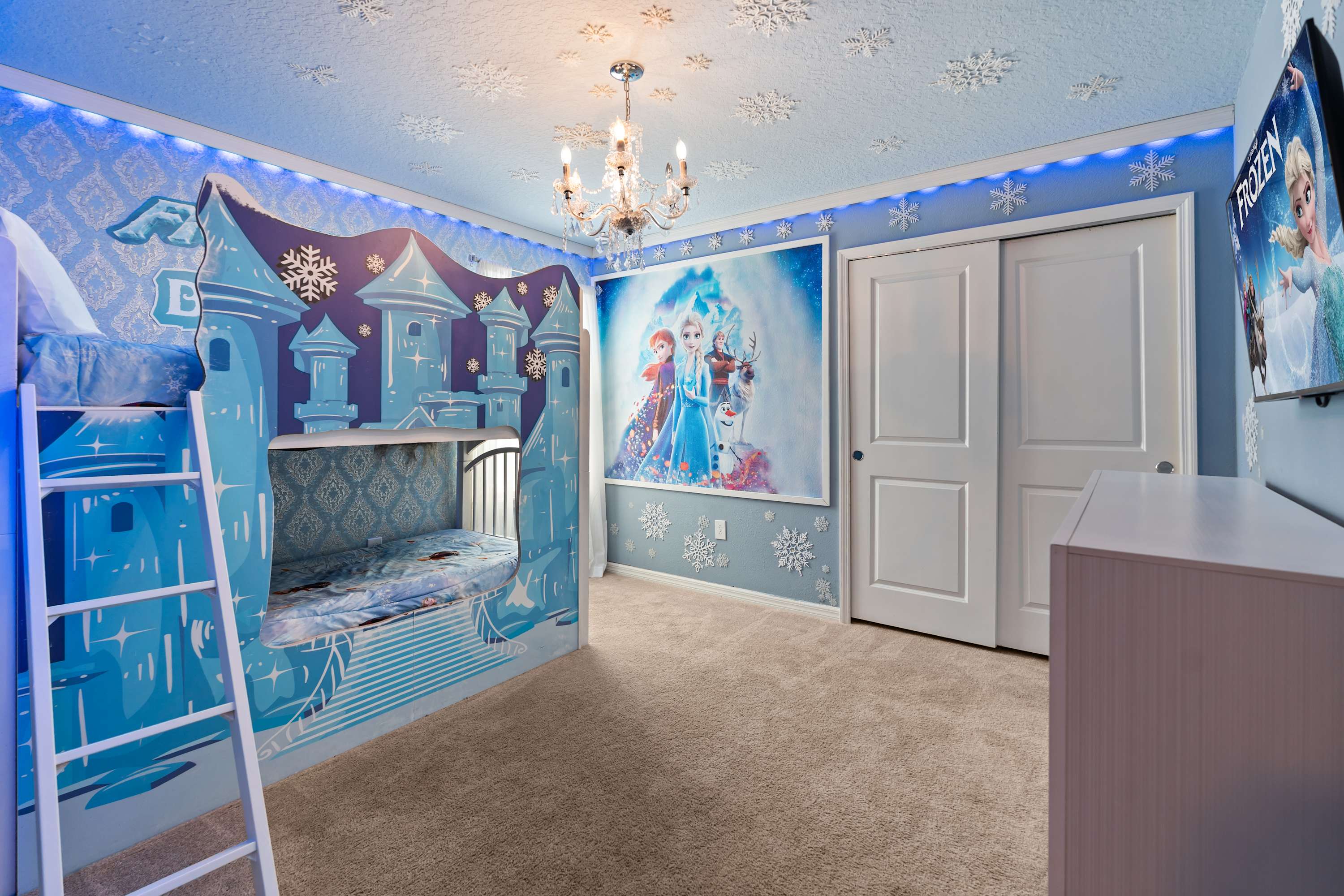 A wider view of the Frozen-themed bedroom shows the castle bunk bed, a large mural of Elsa, Anna, and Olaf, white closet doors, and a wall-mounted TV, all set against light blue walls with snowflake accents.