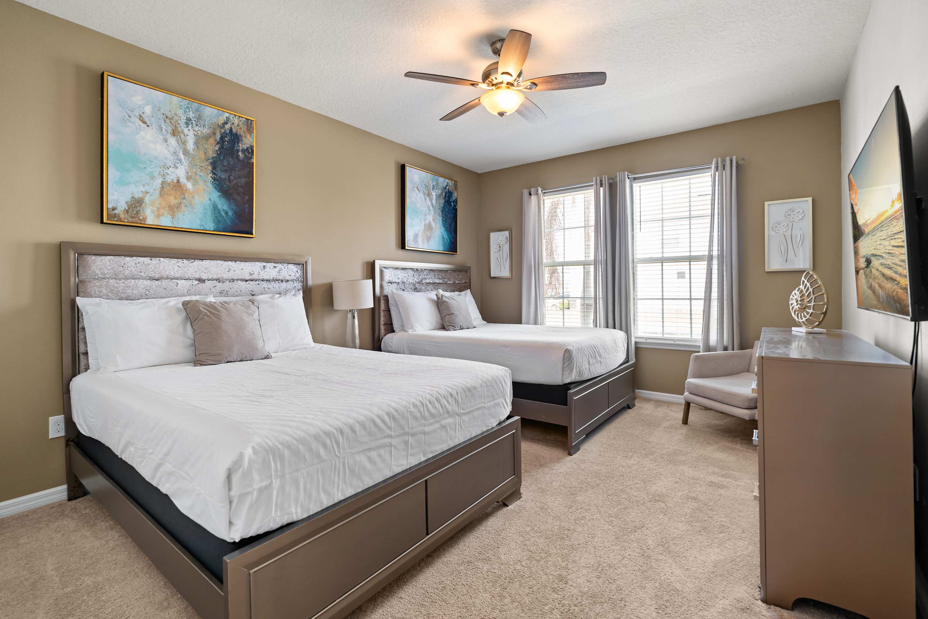 A bright bedroom features two queen-sized beds with white bedding and metallic headboards, set against light brown walls, with a ceiling fan and two windows providing natural light.