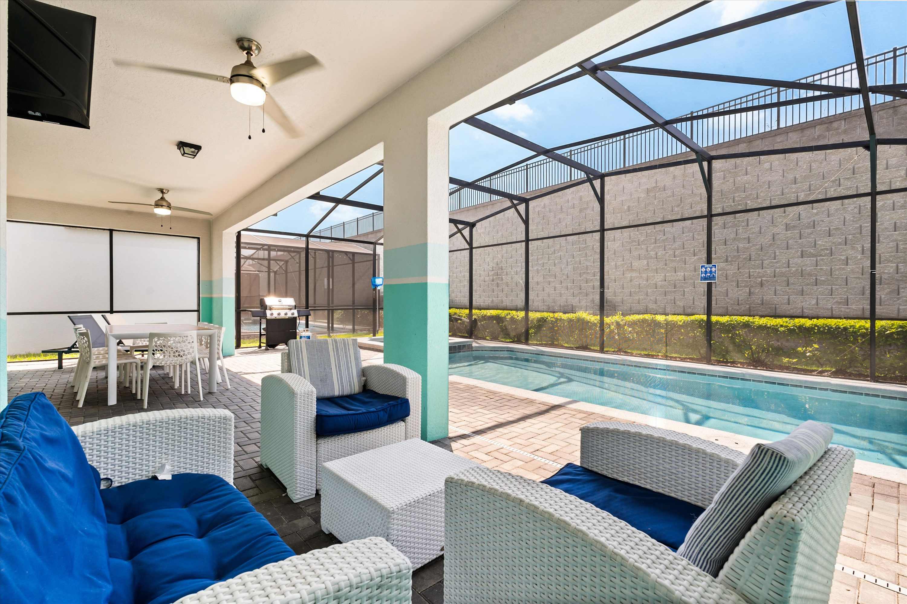 Covered outdoor patio area featuring white wicker seating with blue cushions, a white coffee table, and a view of the screened-in swimming pool, with ceiling fans for comfort.