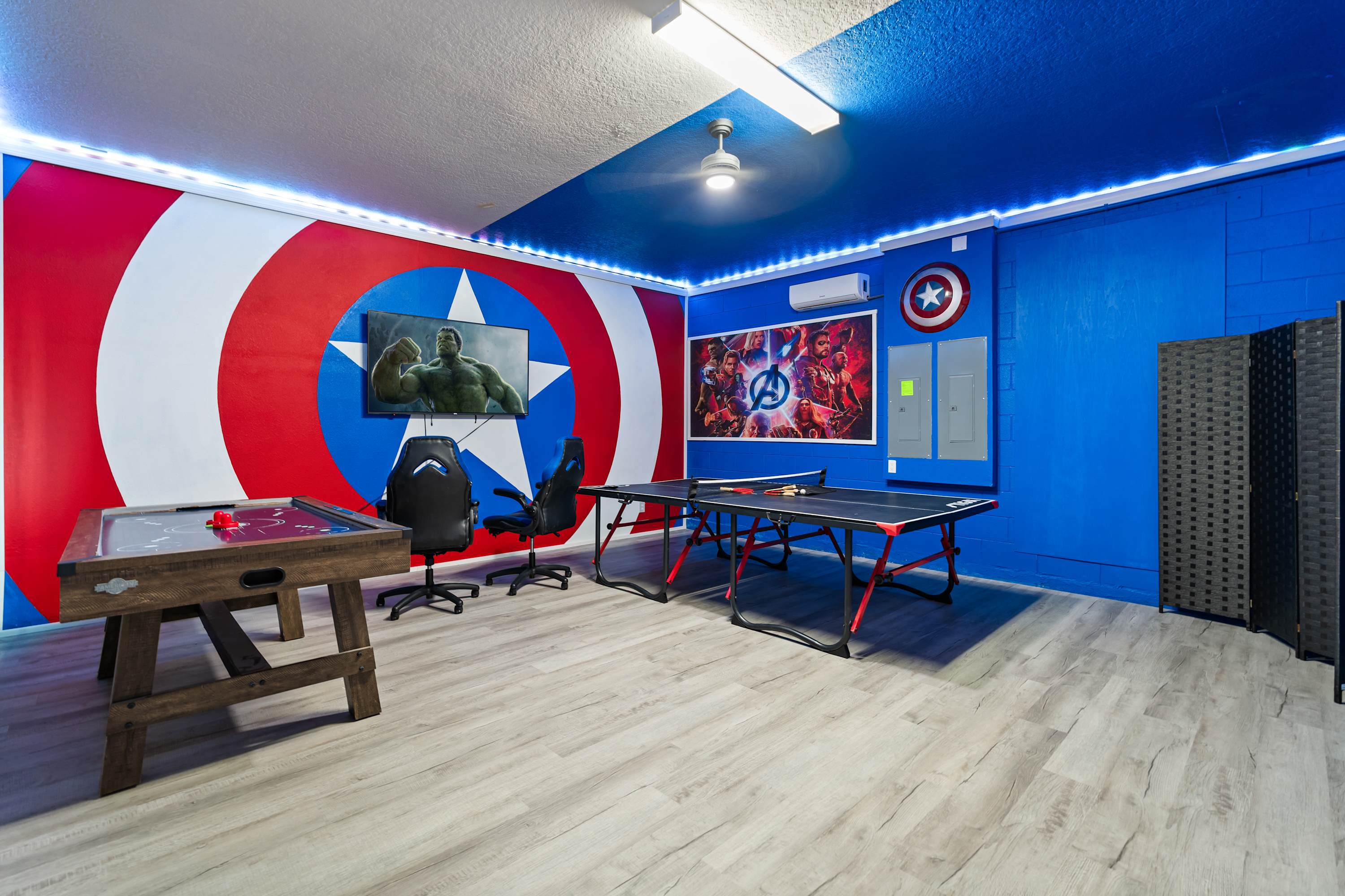 Energetic Marvel-themed game room featuring a large Captain America shield mural with a TV, an air hockey table, and a ping pong table, all set on light wood-look flooring with blue walls and LED strip lighting.