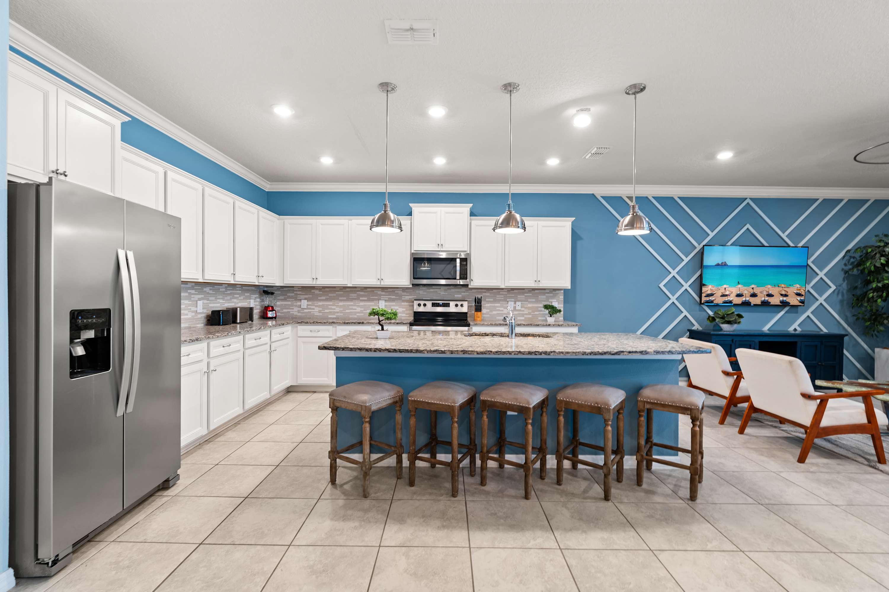 Spacious open-concept kitchen with white cabinets, granite countertops, a large island with four bar stools, and stainless steel appliances, flowing into a living area with a blue accent wall and a wall-mounted TV.