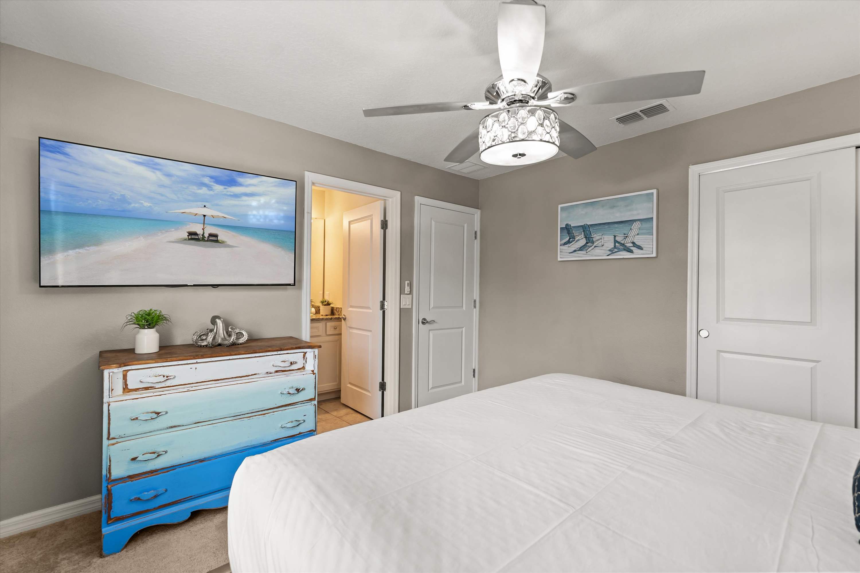 A view of the bedroom showing a large TV mounted above a distressed blue dresser, with a white bed in the foreground. A door leads to an ensuite bathroom, and two white closet doors are visible on the right.