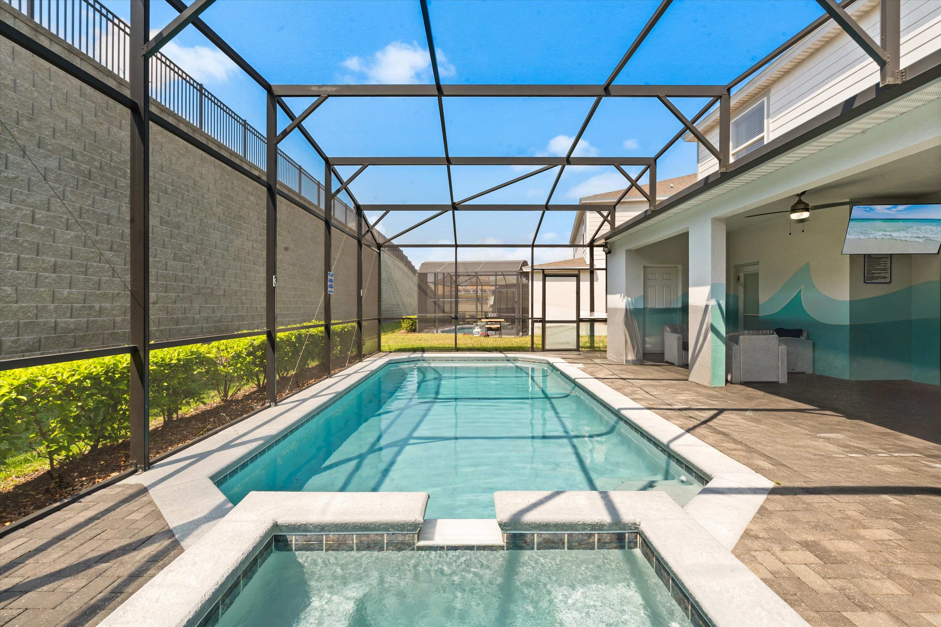 Wide view of the screened-in outdoor pool and hot tub area, surrounded by a paved deck and a tall privacy wall, with clear blue water reflecting the sunny sky.