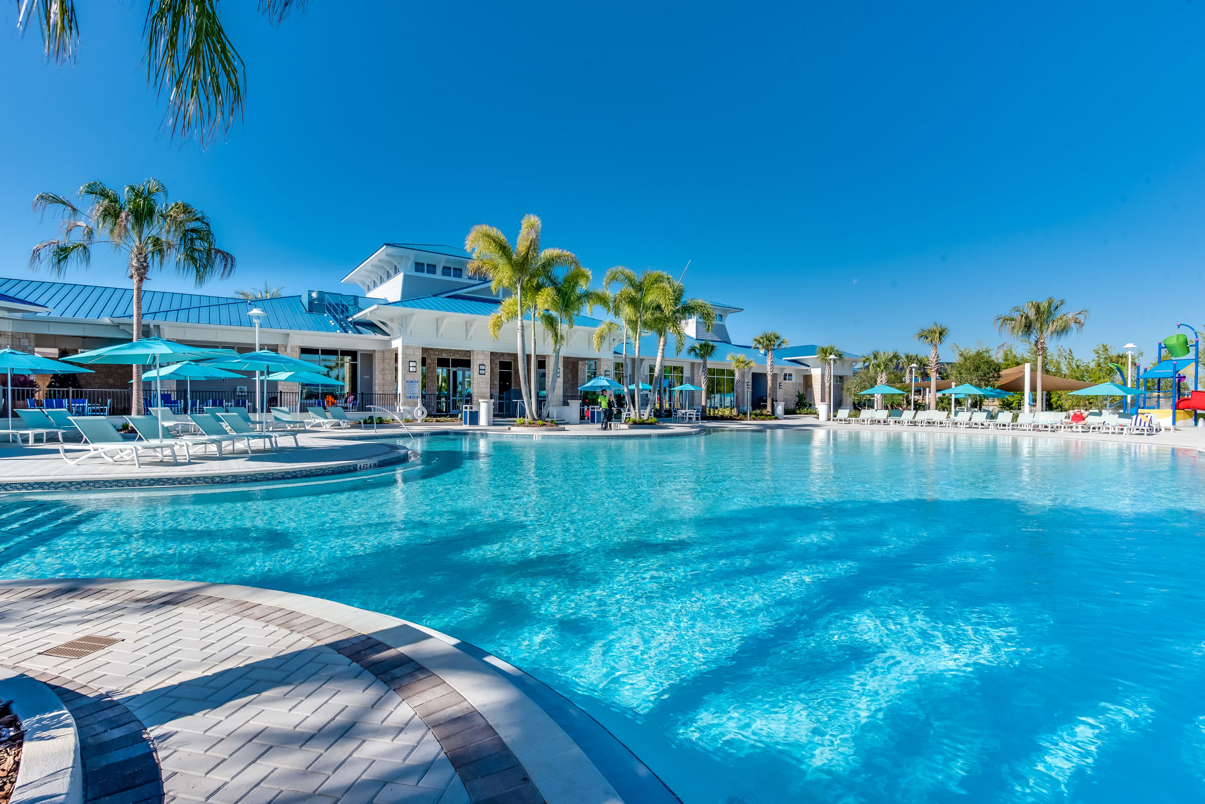 A ground-level view captures the sparkling blue waters of a large communal swimming pool, surrounded by lounge chairs with blue umbrellas and a modern clubhouse in the background under a clear sky.