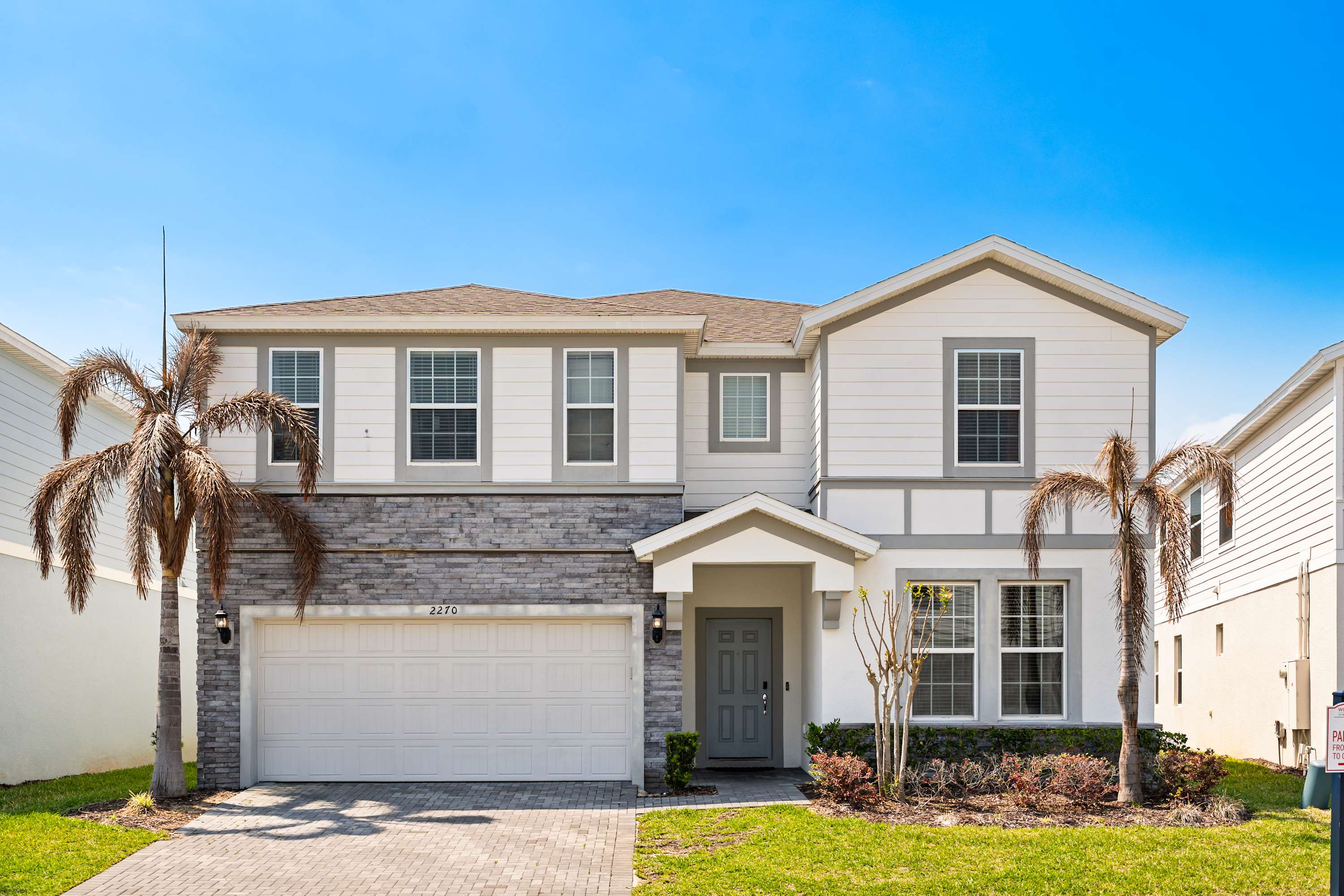 A two-story modern house with a light-colored facade, grey stone accents, a two-car garage, and a grey front door, flanked by palm trees and a green lawn under a clear blue sky.